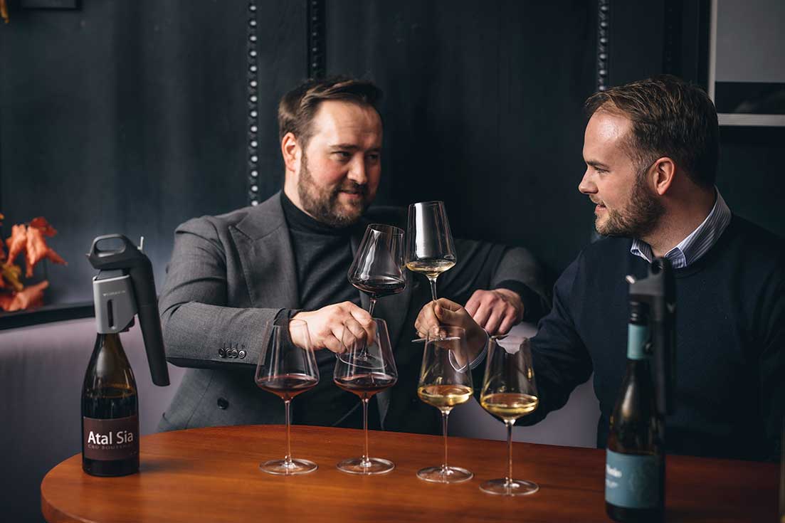 Two men clinking glasses of red and white wine at a wooden table, with Coravin wine preservation systems attached to opened wine bottles.