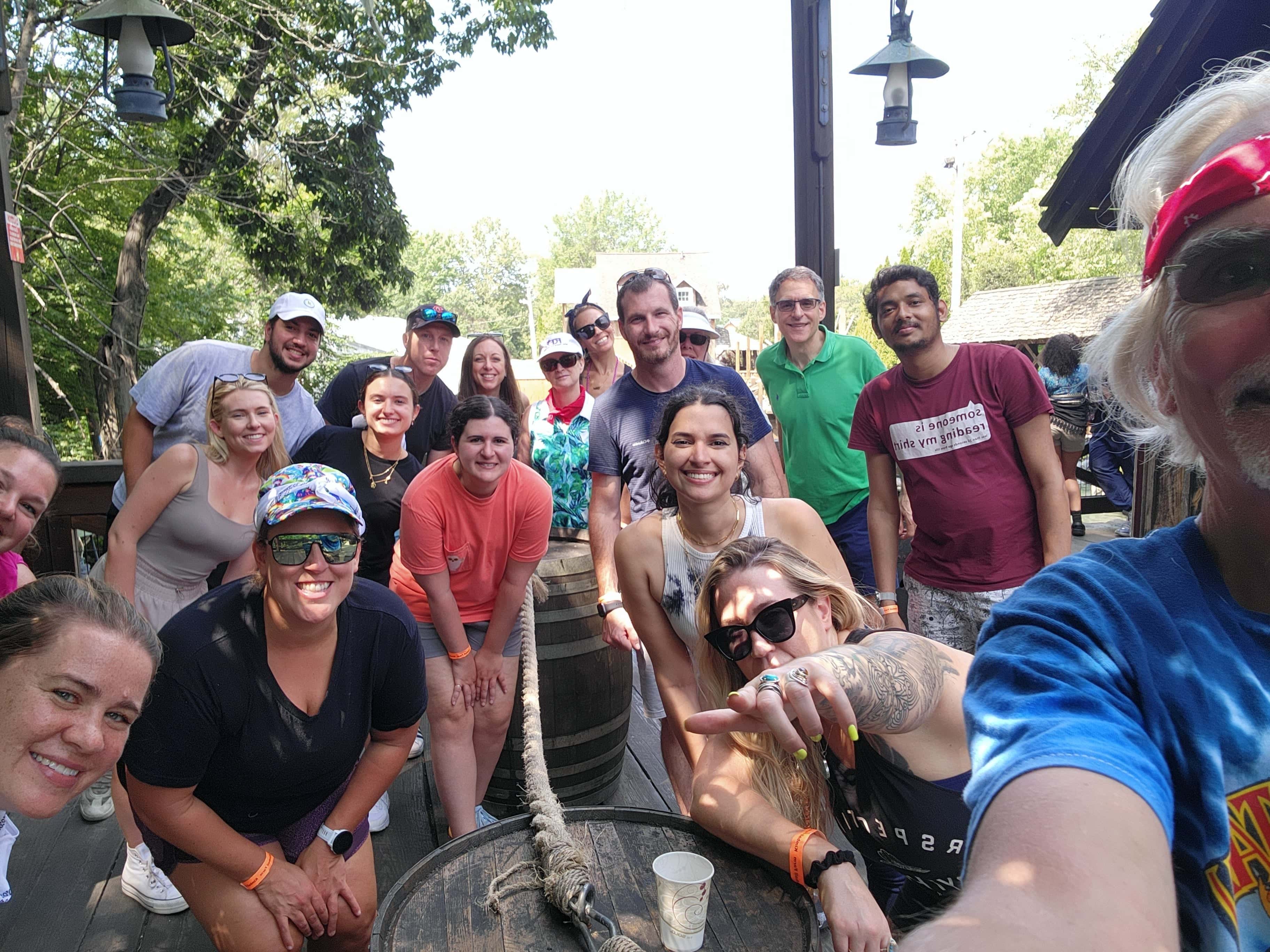 Group of smiling people gathered around a wooden barrel, enjoying a sunny day outdoors.