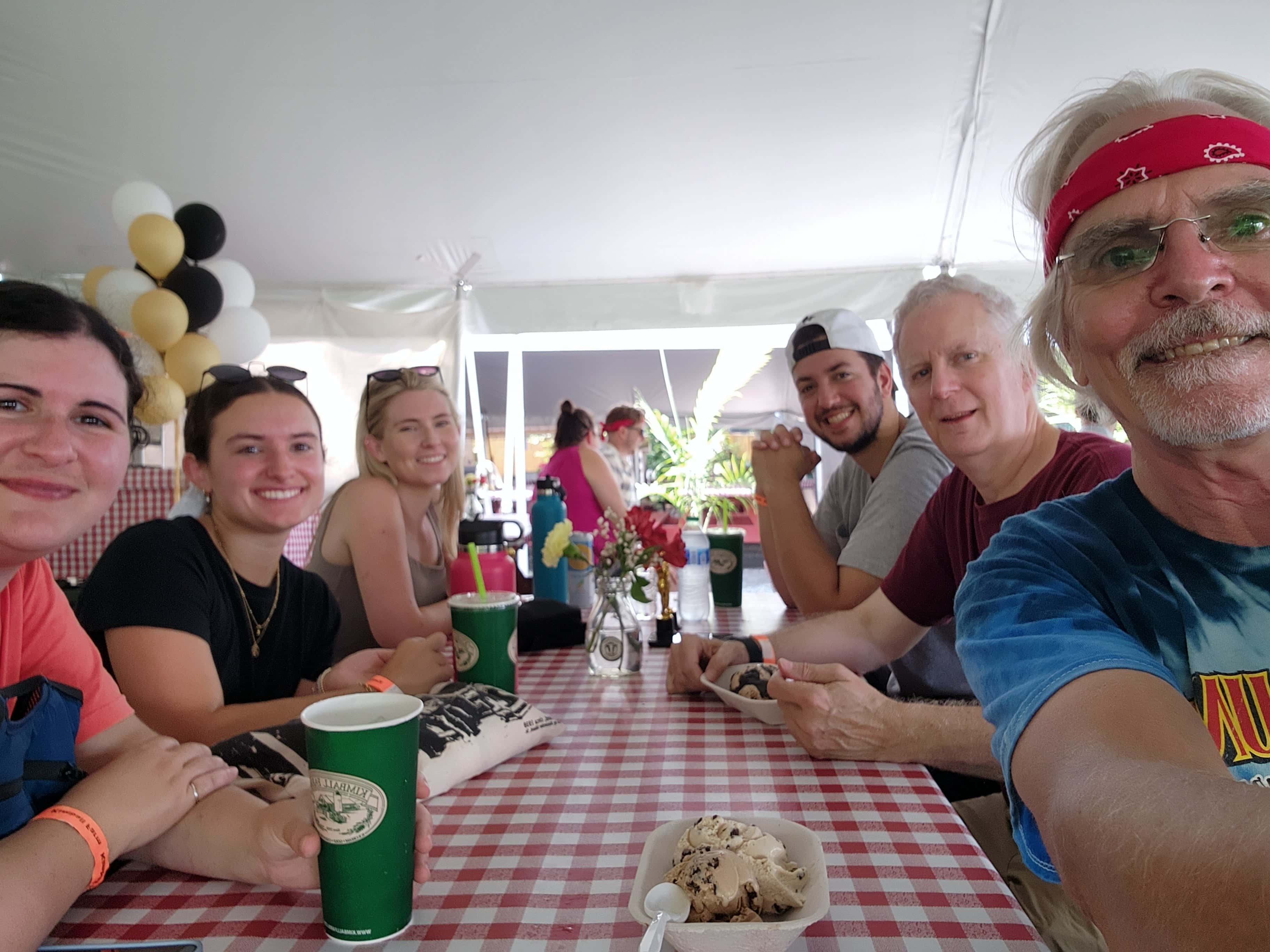 Group of five people smiling at a table with food, drinks, and flowers, under a tent decorated with balloons.