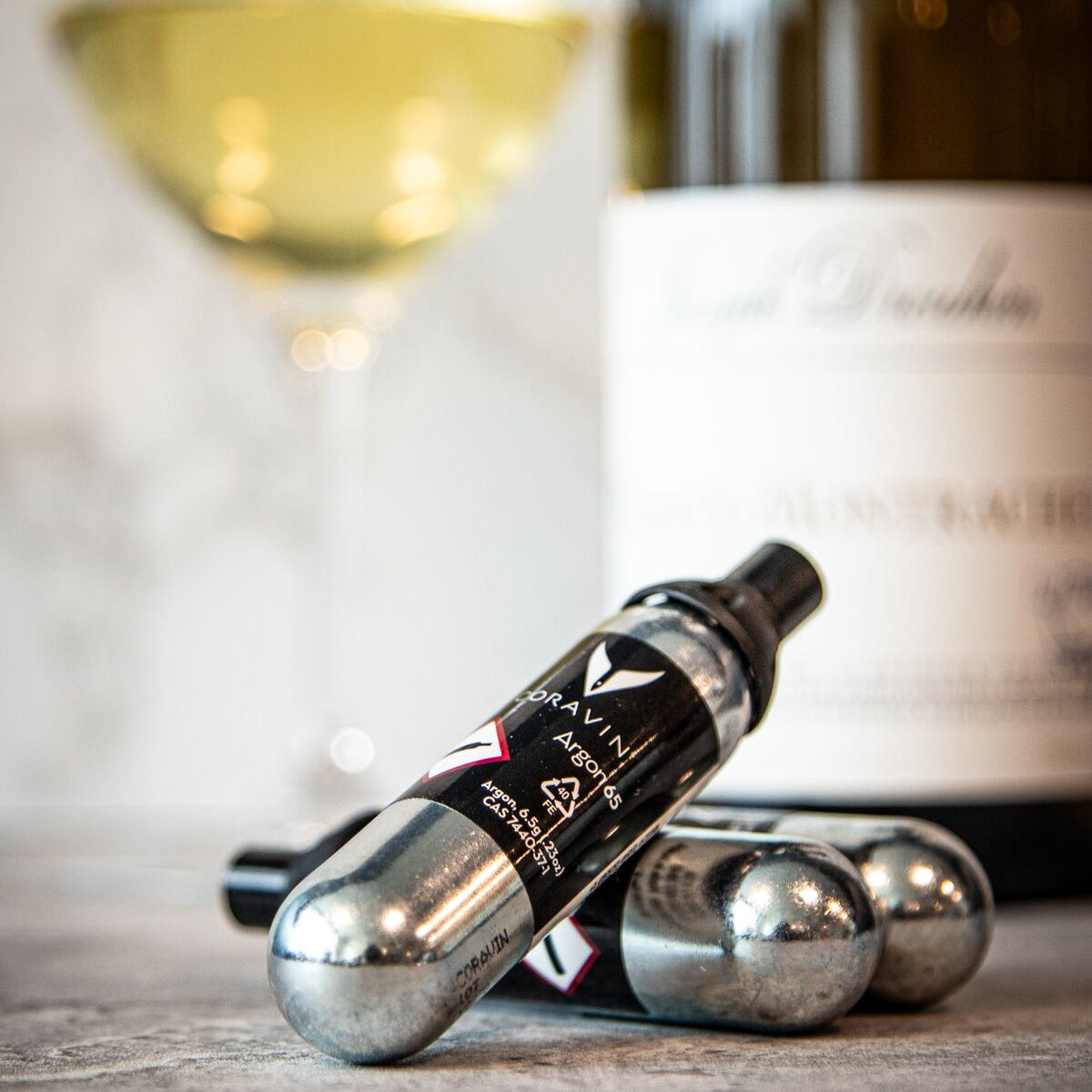 A glass of white wine with a bottle in the background and two silver gas canisters labeled "Coravin."