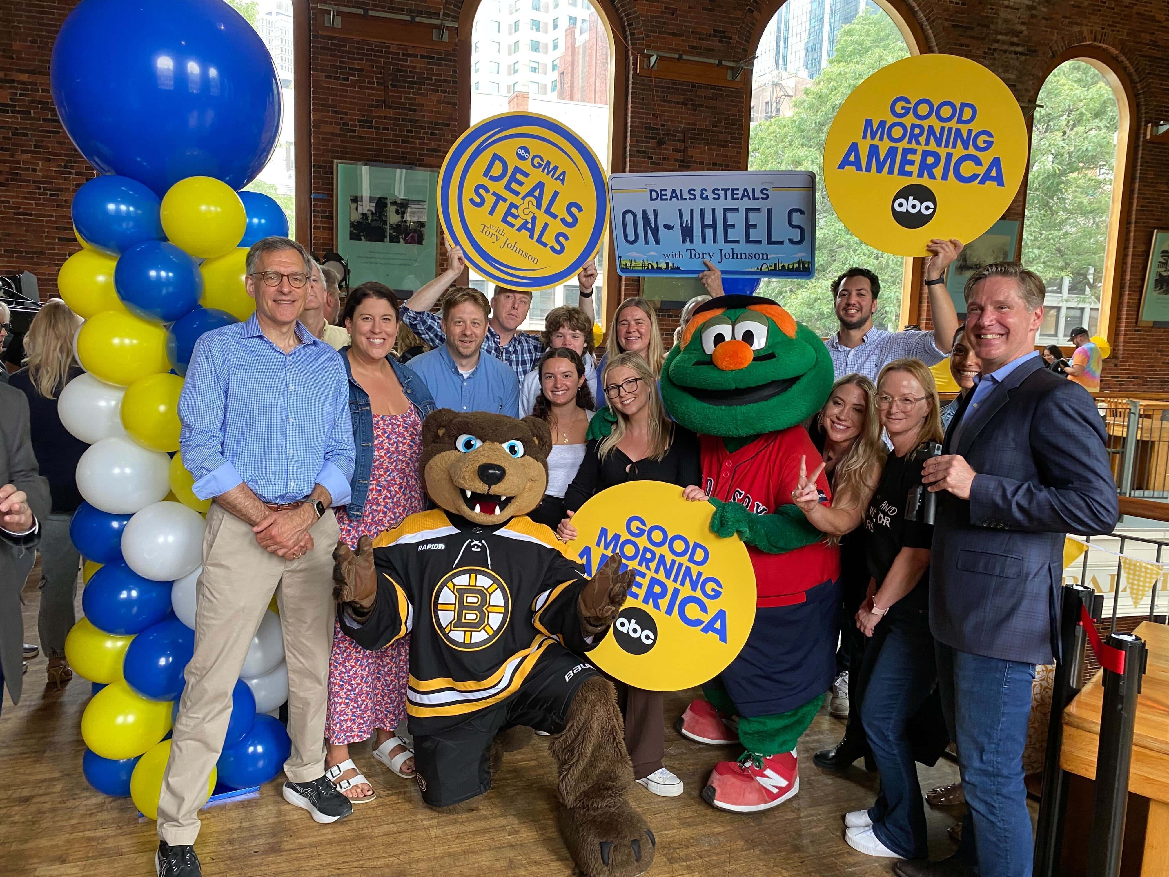 Group photo featuring hosts and mascots holding signs for "Good Morning America" and "Deals & Steals," with colorful balloons in the background.