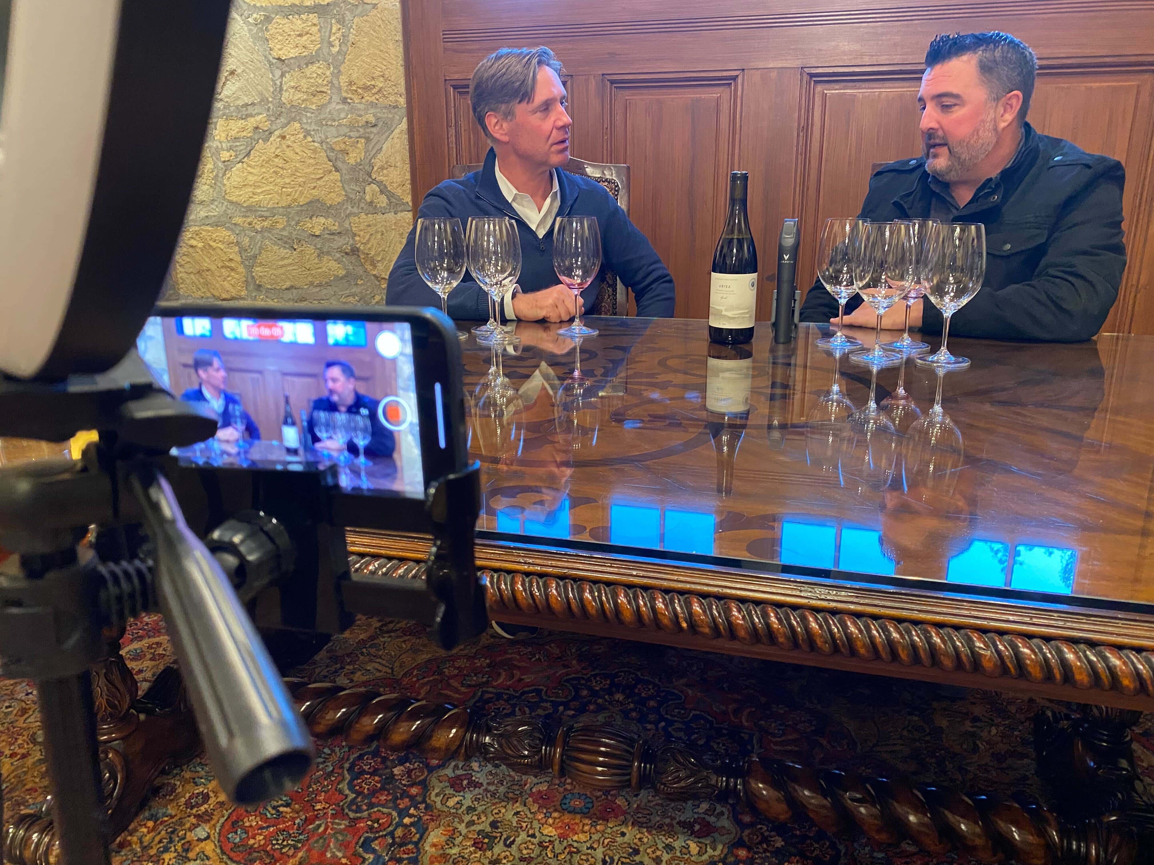 Two men engaged in conversation over a wooden table with wine glasses and a bottle, filmed by a smartphone on a tripod.