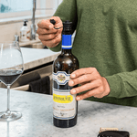 Hand opening a bottle of Rombauer Cabernet Sauvignon next to a glass of red wine on a kitchen countertop.