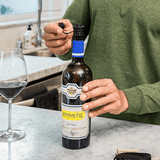 Hand opening a bottle of Rombauer Cabernet Sauvignon next to a glass of red wine on a kitchen countertop.