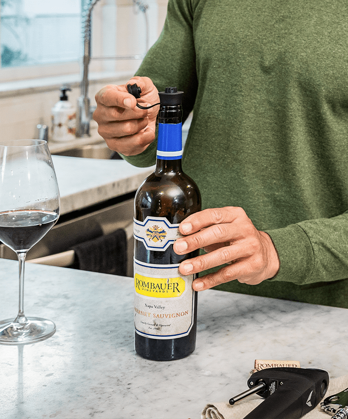 Hand opening a bottle of Rombauer Cabernet Sauvignon next to a glass of red wine on a kitchen countertop.