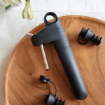 Coravin wine opener in black with two black bottle stoppers on a wooden serving platter, surrounded by flowers.