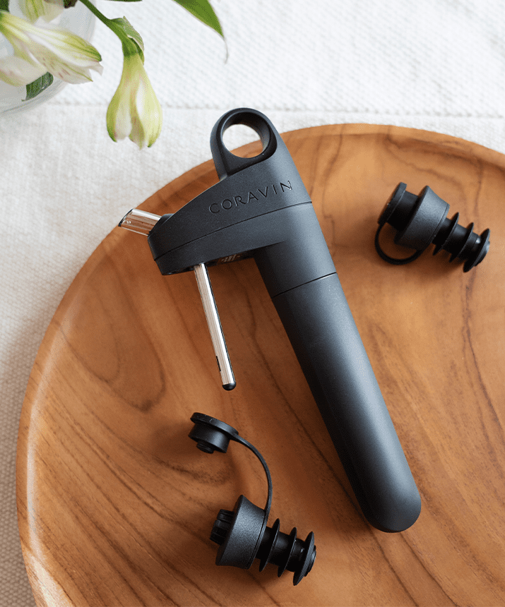 Coravin wine opener in black with two black bottle stoppers on a wooden serving platter, surrounded by flowers.