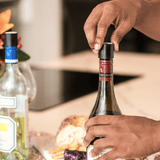A hand removes the cork from a red wine bottle with two other bottles and a cheese platter in the background.