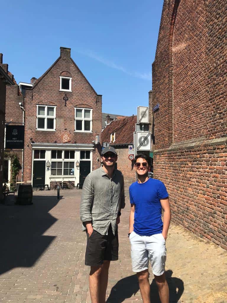 Two men stand on a cobblestone street near brick buildings, enjoying a sunny day in a quaint town.