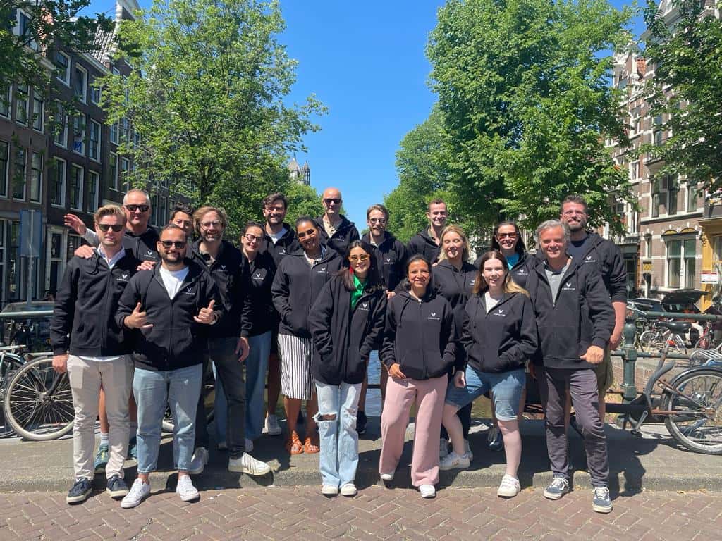 Group of diverse individuals in black jackets posing together on a sunny day in Amsterdam, surrounded by trees and bicycles.