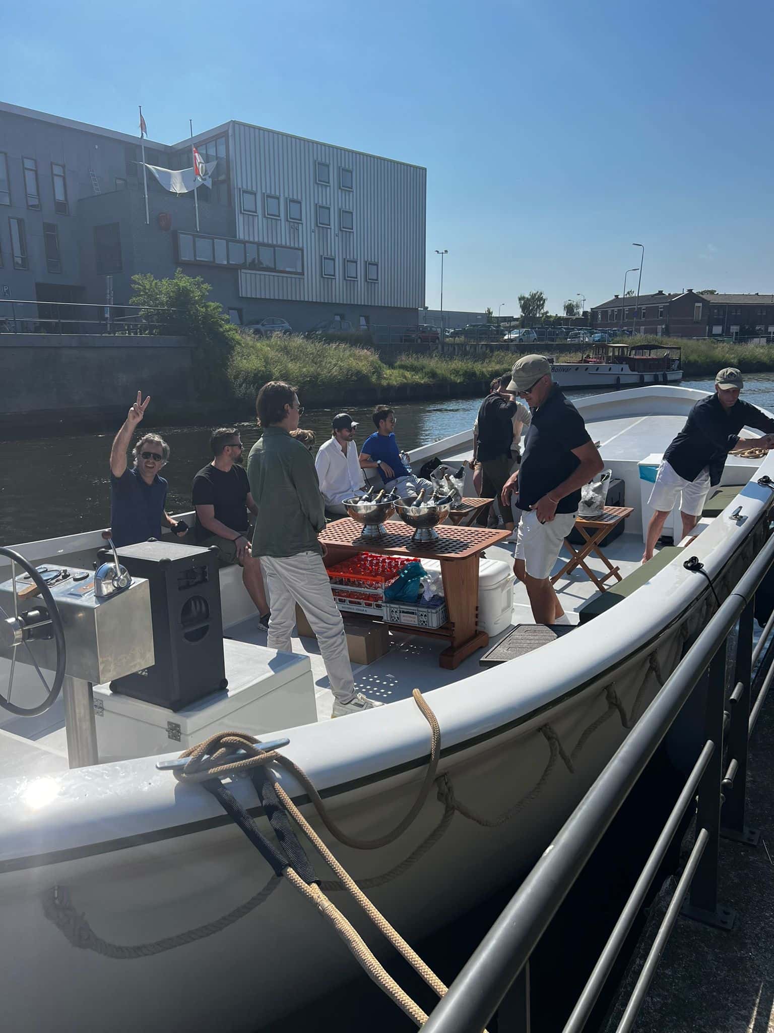 Group of people on a boat, some socializing, with a building and flags visible in the background under a clear blue sky.