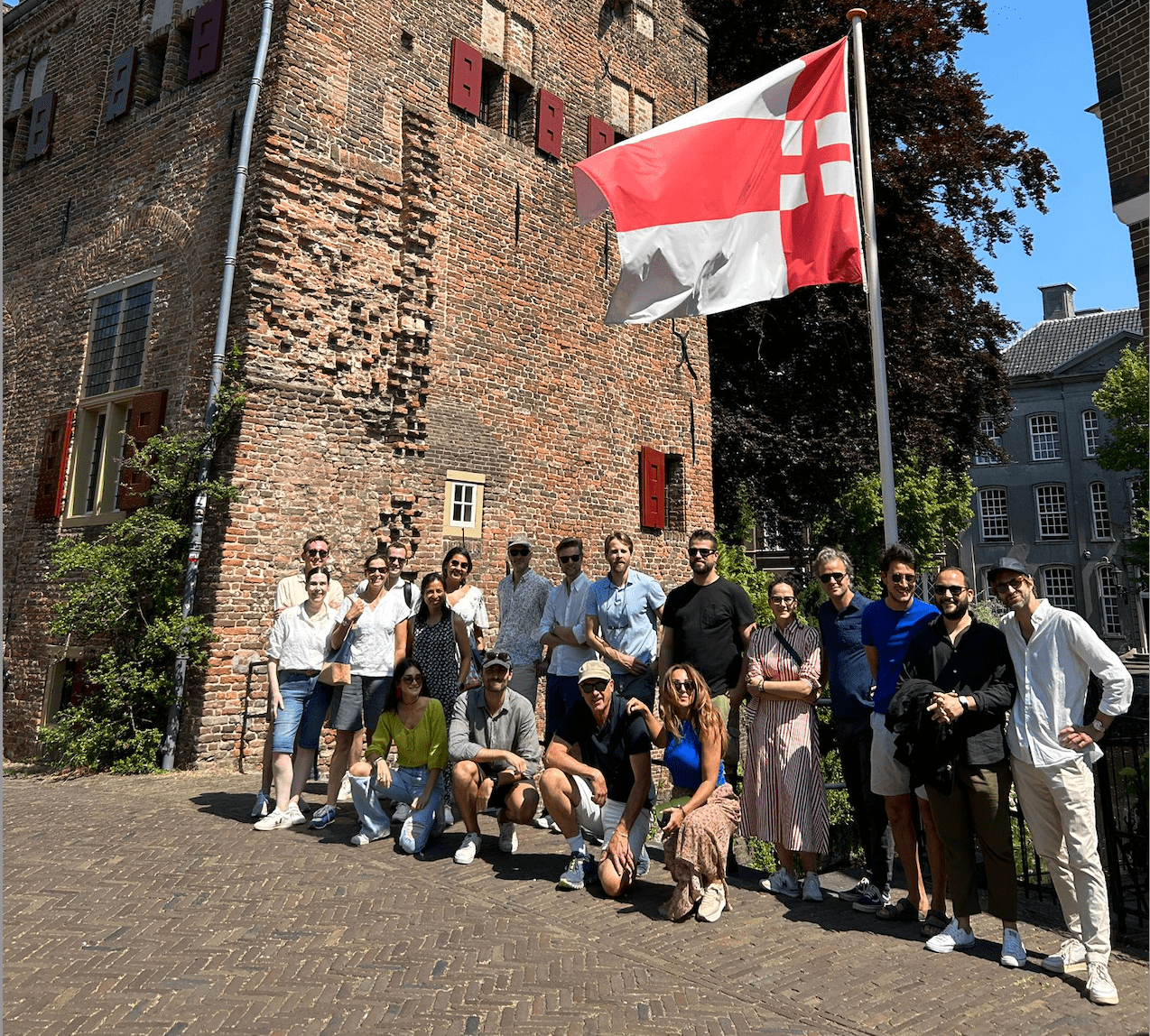 Group of people posing in front of a brick building with red shutters and a flag featuring a red cross on a white background.