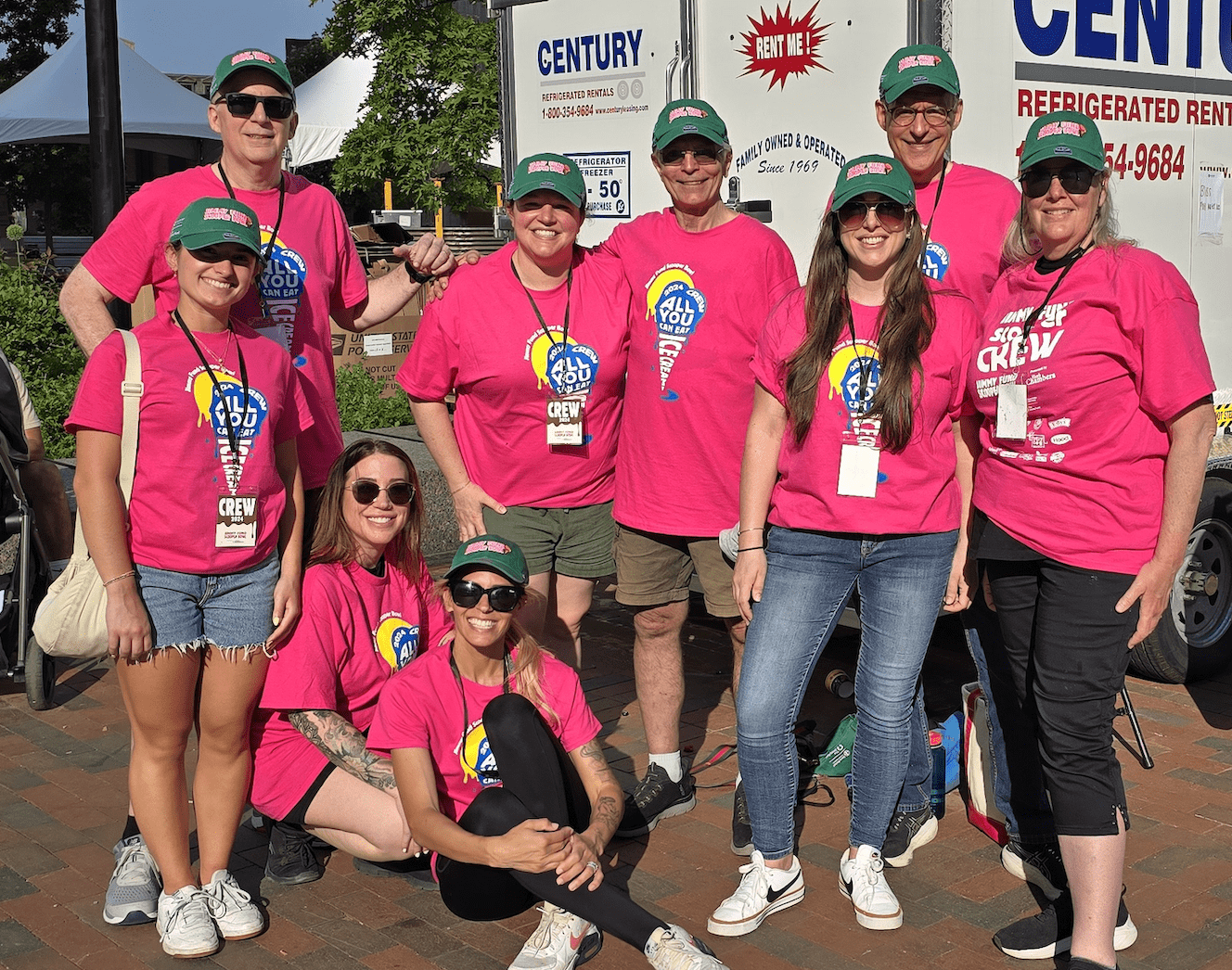 Group of ten people in matching pink shirts and green hats, posing together at an outdoor event.