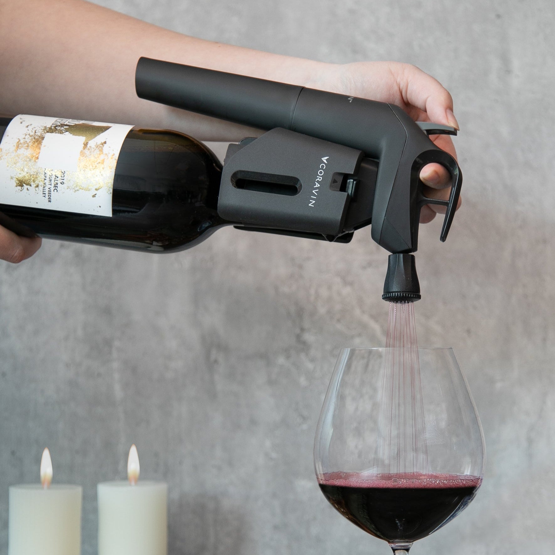 Hand pouring red wine from a bottle using a Coravin wine preservation system into a glass beside lit candles.
