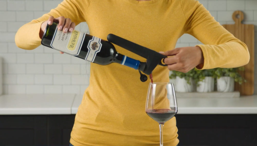 A person in a yellow sweater pours red wine from a bottle into a glass using a wine aerator.