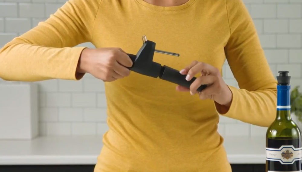 A person in a yellow shirt uses a wine opener over a bottle of wine on a kitchen counter.