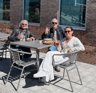Three people enjoying drinks at an outdoor café table, with sunlight and a modern building in the background.
