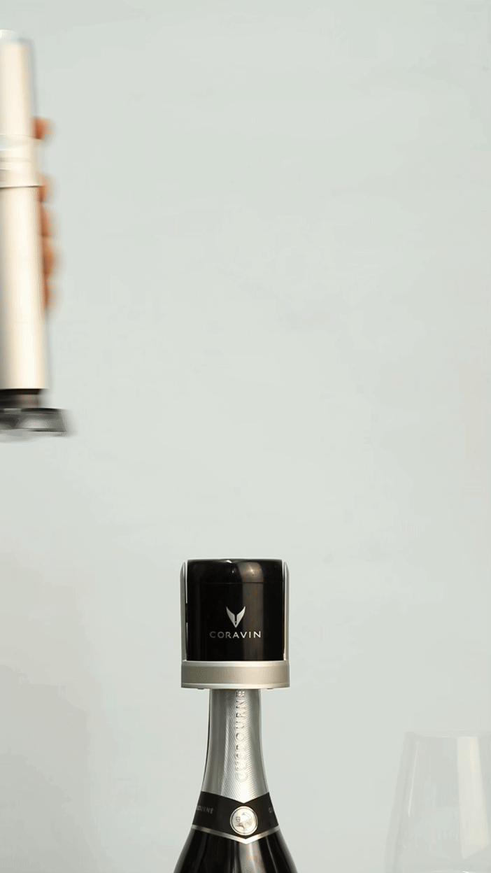 A hand prepares to use a Coravin wine preservation system on top of a bottle, with a wine glass nearby.