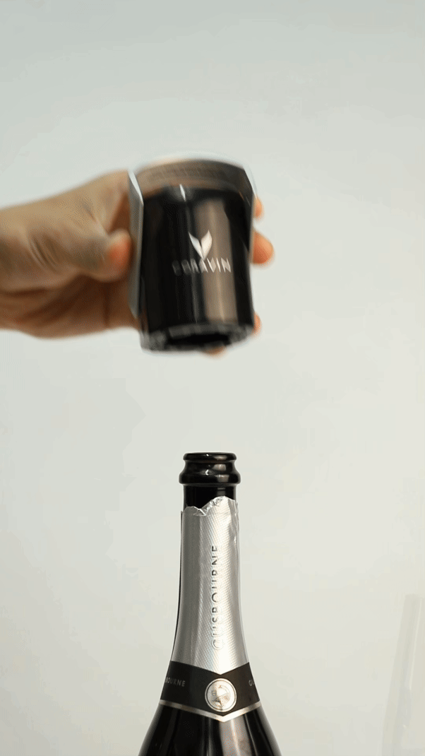 A hand holds a Coravin device above a champagne bottle, preparing to extract wine without removing the cork.