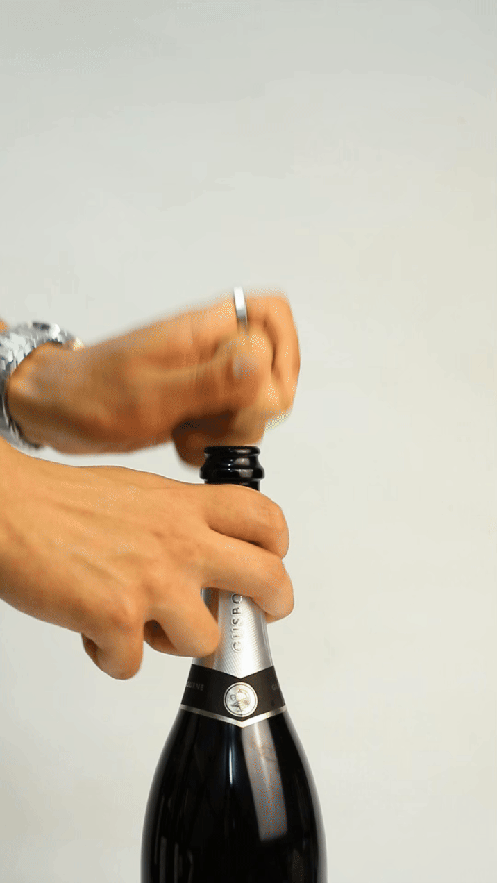 Hands gripping the neck of a sparkling wine bottle, twisting the cork to open it, with a silver cap visible.