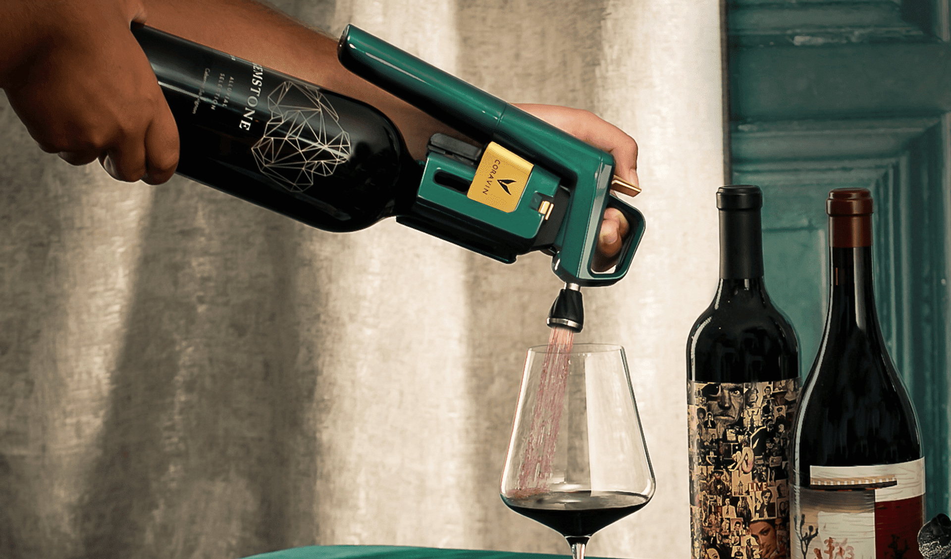Hand pouring red wine using a green and gold Coravin wine preservation system into a glass, with two additional wine bottles standing nearby on a teal-colored table.