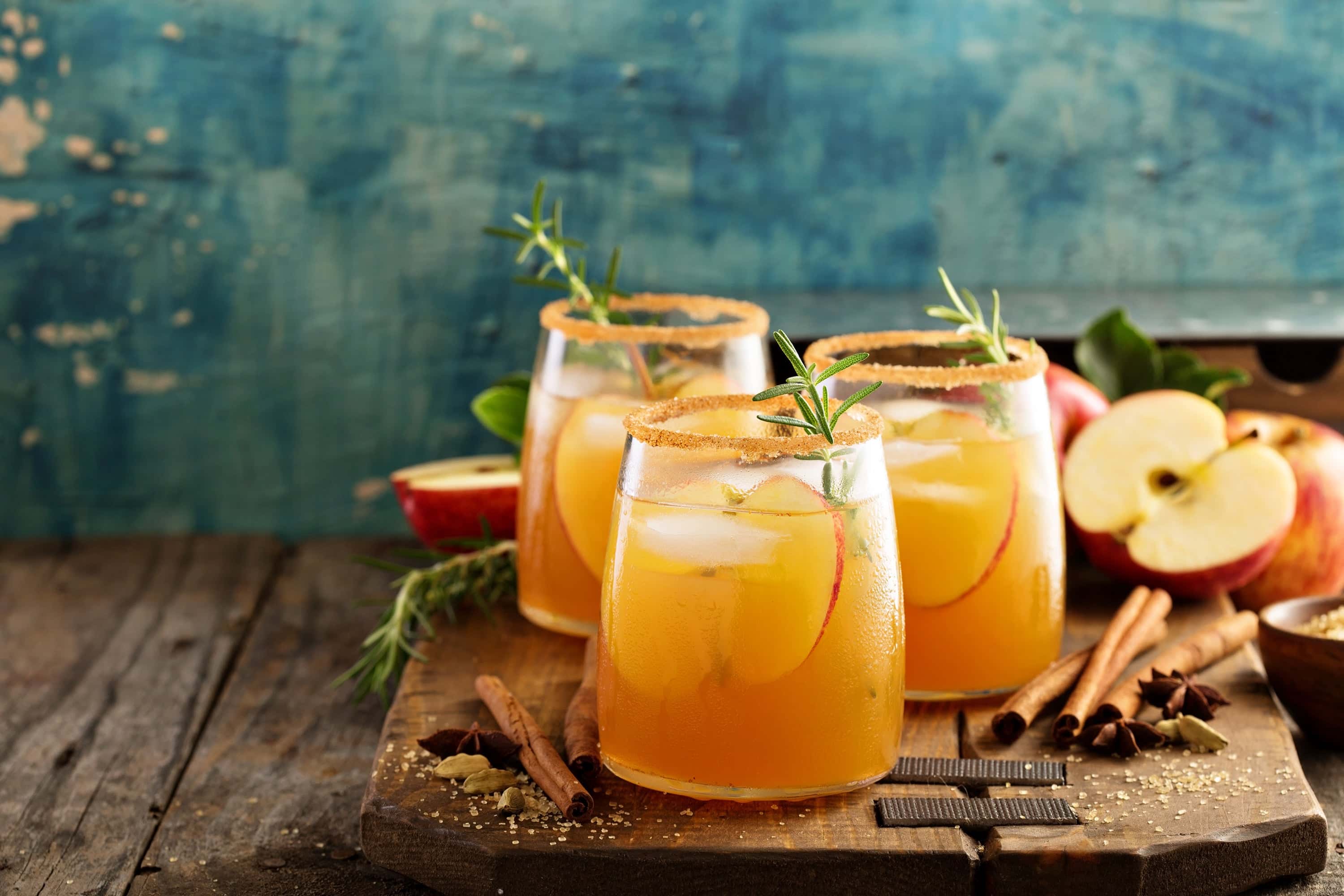 Three glasses of apple cider cocktails with cinnamon sugar rims, garnished with apple slices and rosemary, surrounded by fresh apples, cinnamon sticks, and spices on a rustic wooden board.