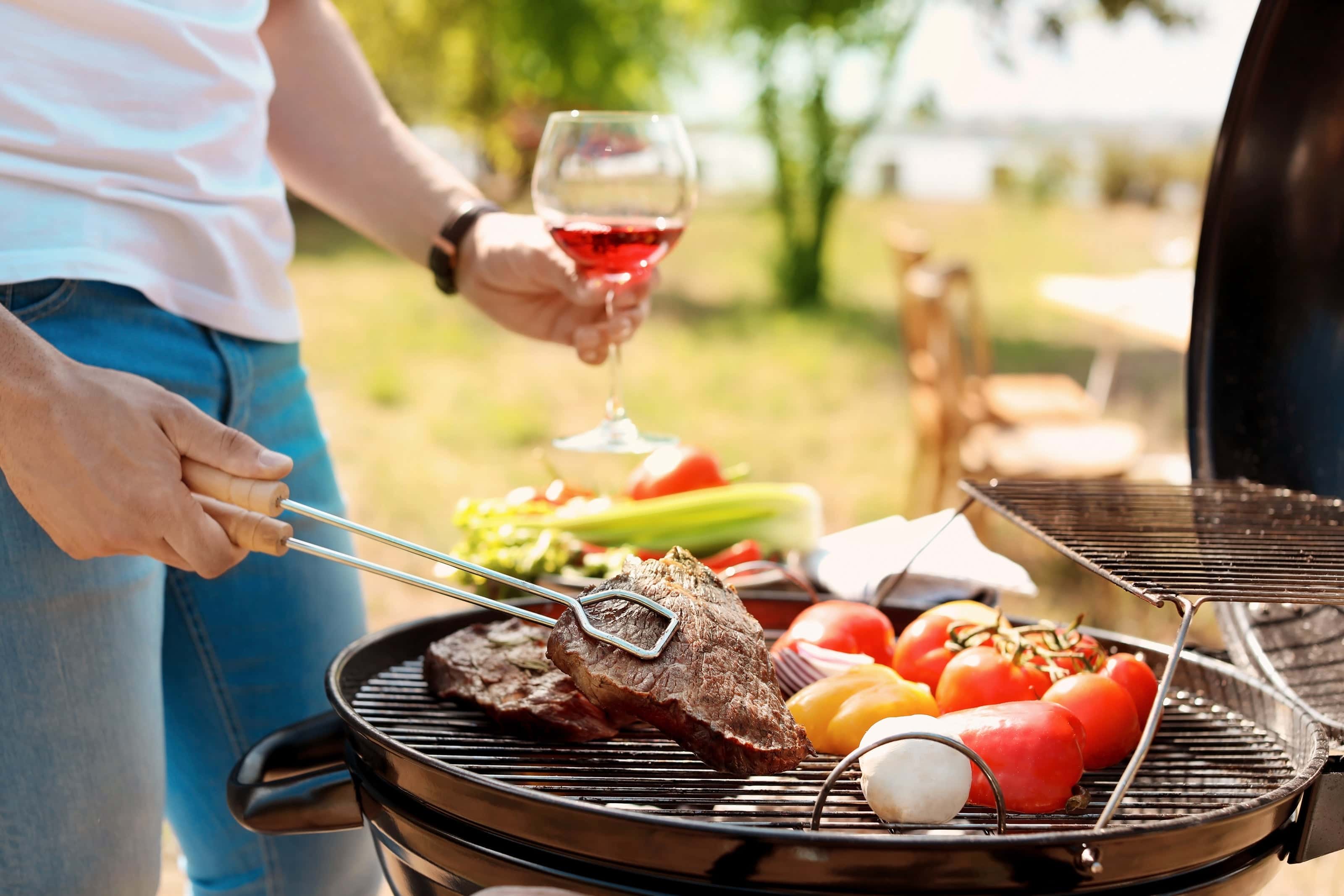 Person grilling steak and vegetables on a charcoal barbecue while holding a glass of rosé wine outdoors on a sunny day.