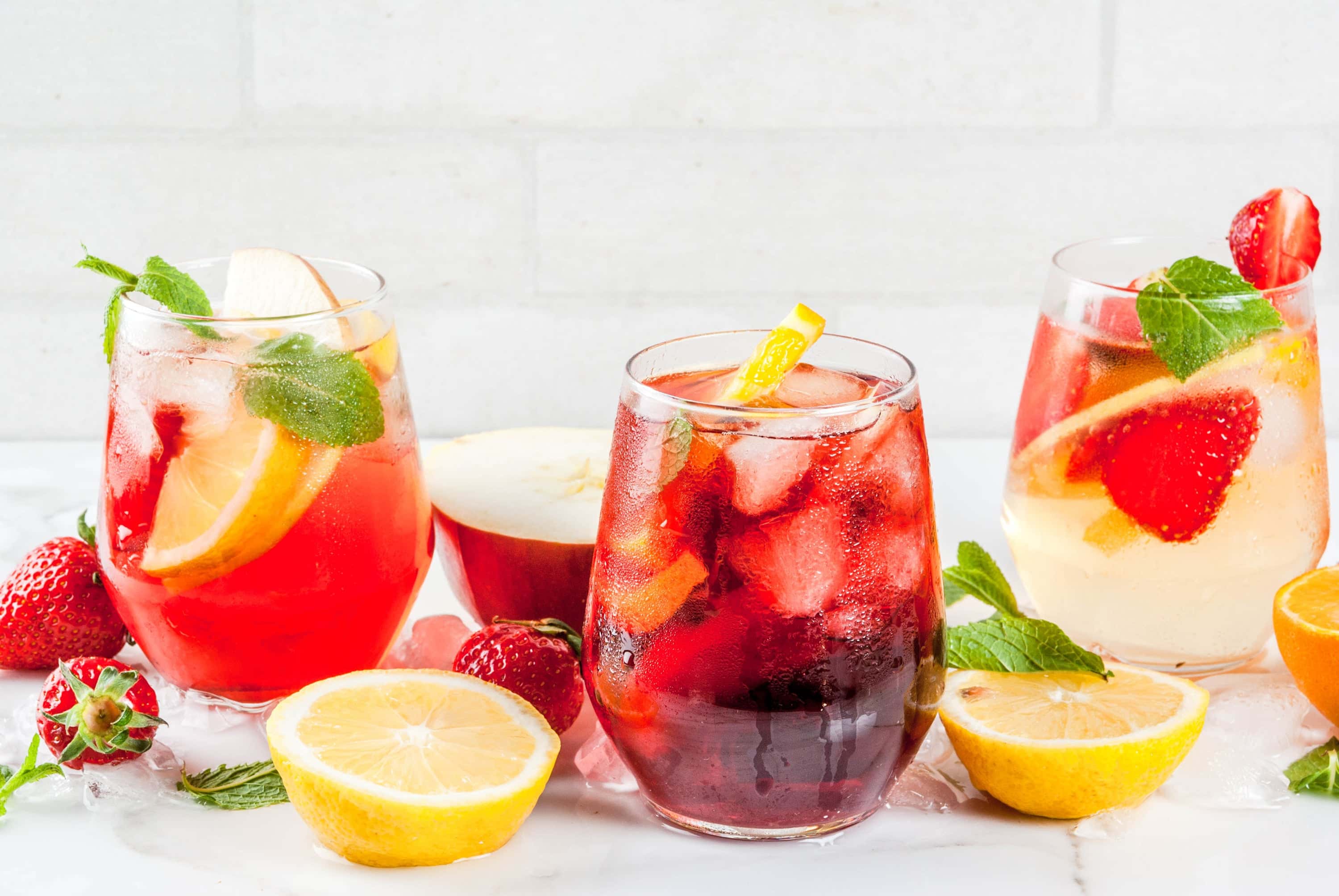 Three glasses of fruity sangria garnished with lemon, apple, strawberry, and mint, served over ice with fresh ingredients scattered on a white marble surface.
