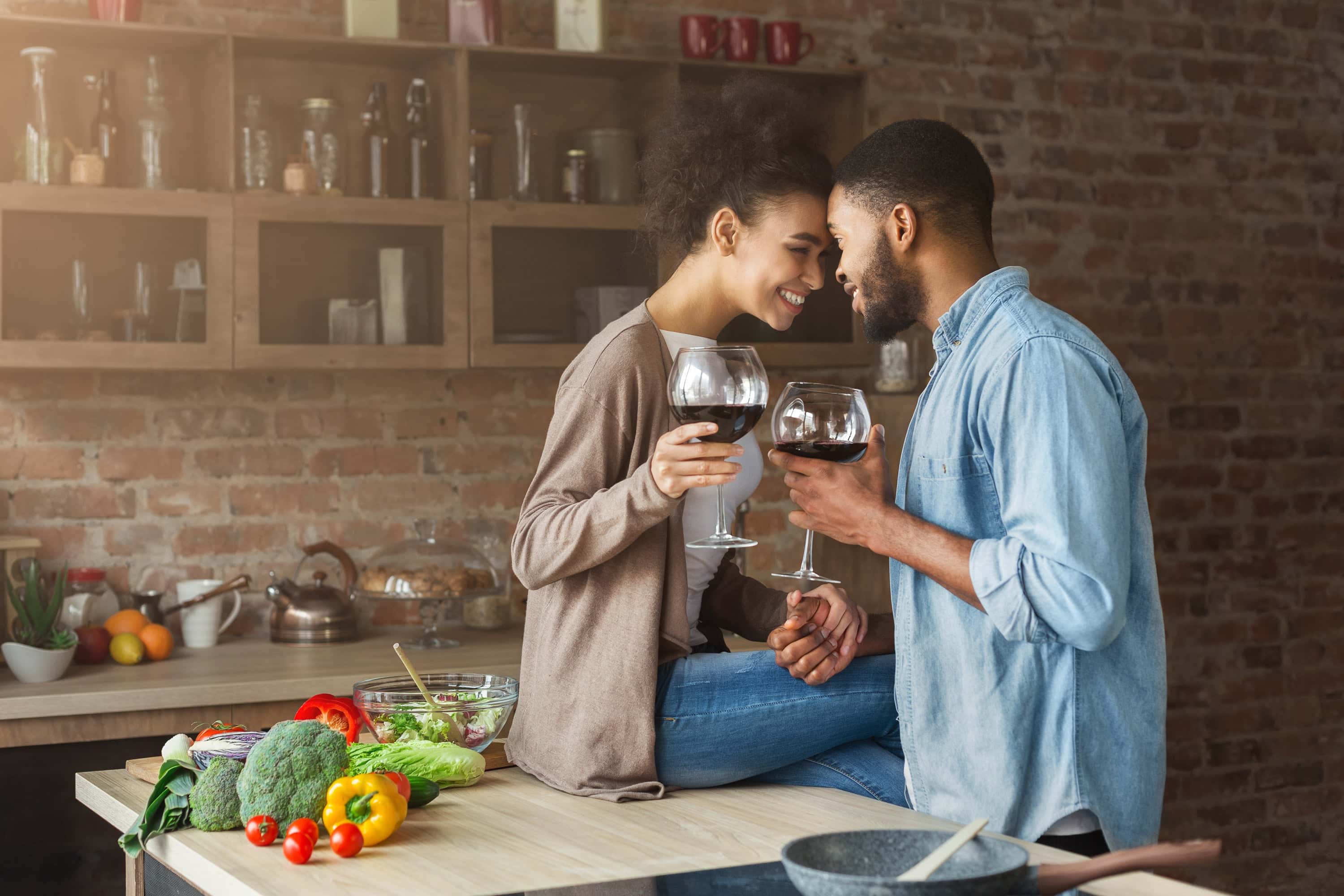 Happy couple in a cozy kitchen toasting with red wine while preparing fresh vegetables together.