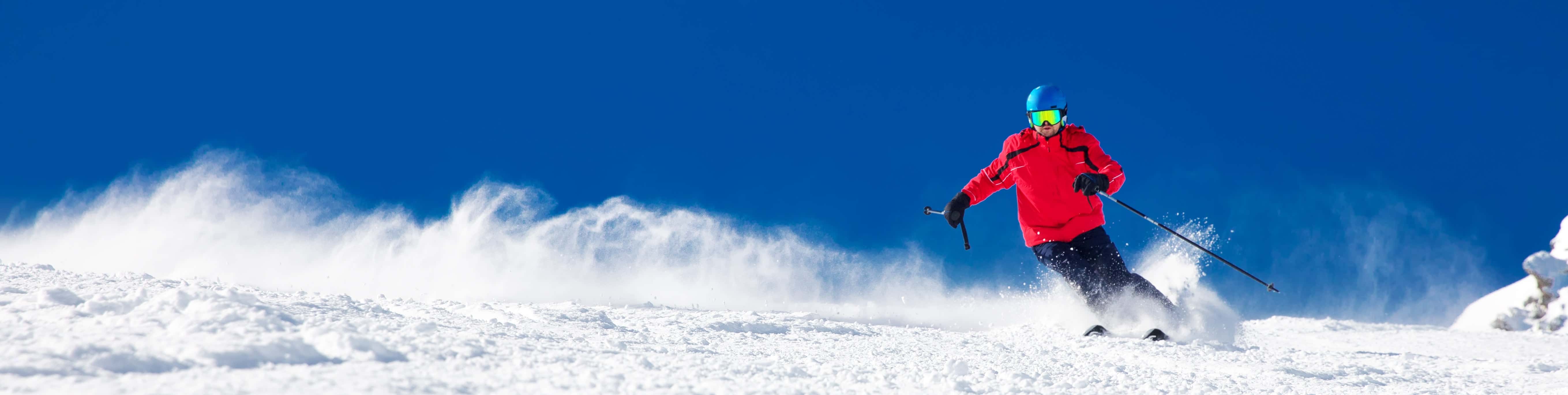 Skier in a red jacket carving down a snowy slope, kicking up powder against a clear blue sky on a bright winter day.