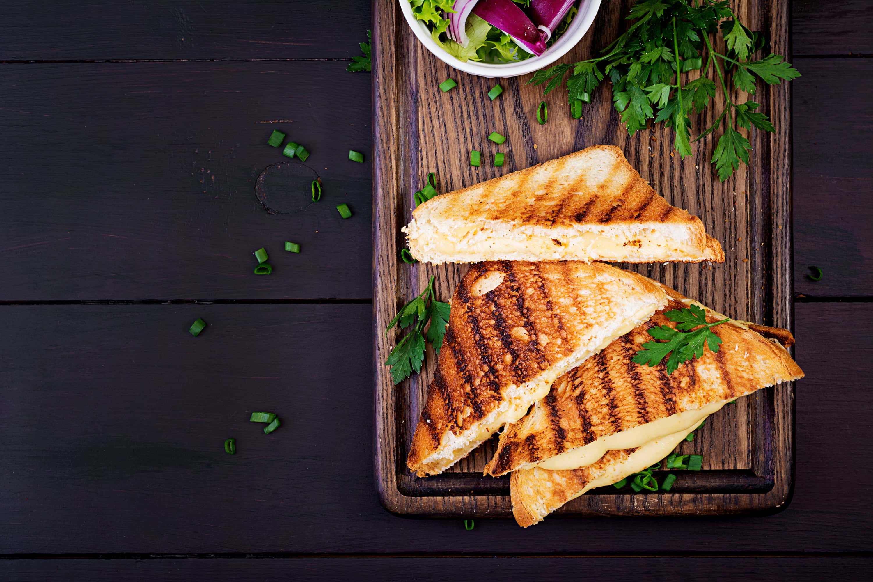 Grilled cheese sandwiches cut into triangles served on a wooden board with fresh parsley and a small side salad, garnished with chopped green onions.