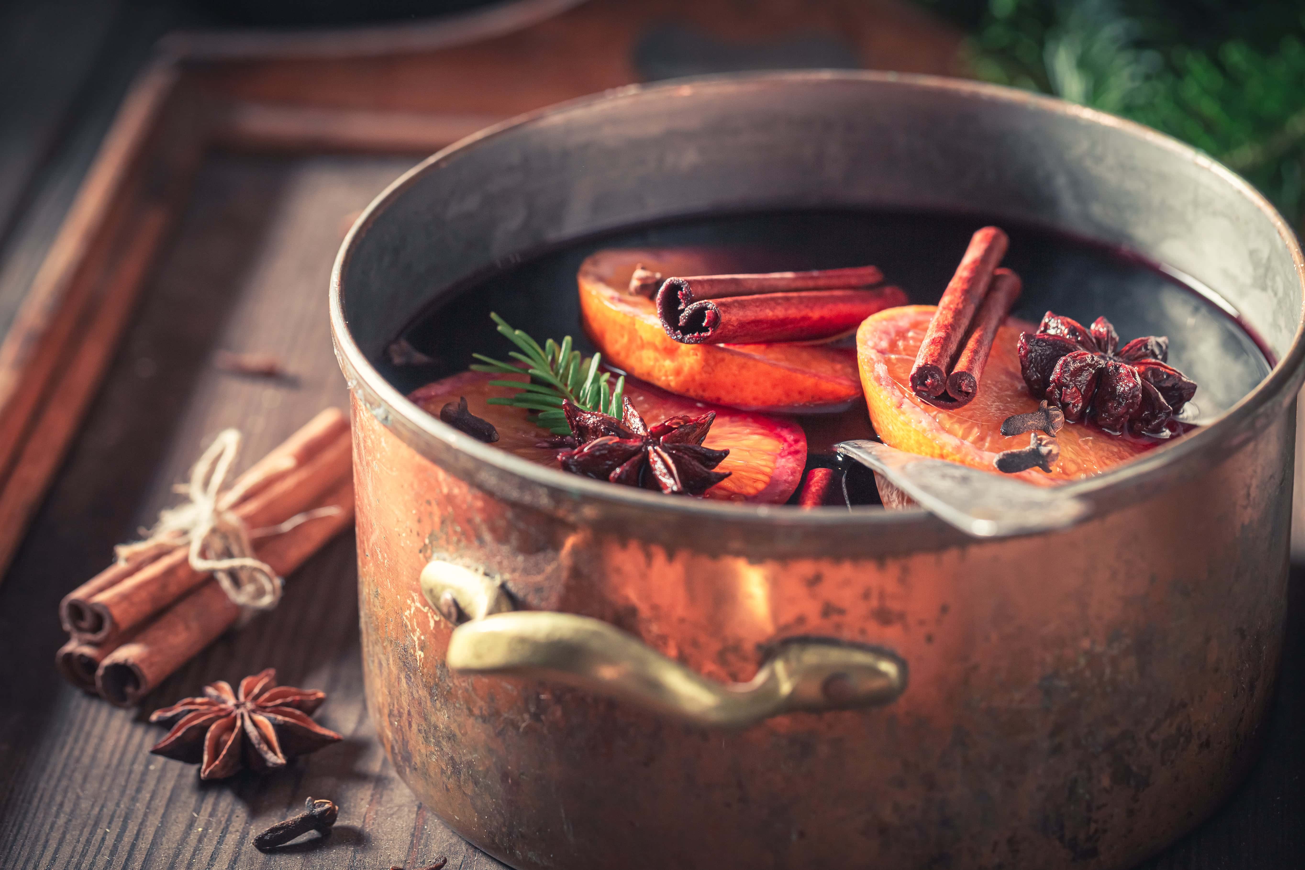 Traditional mulled wine simmering in a rustic copper pot with orange slices, cinnamon sticks, star anise, cloves, and rosemary, set on a wooden surface.