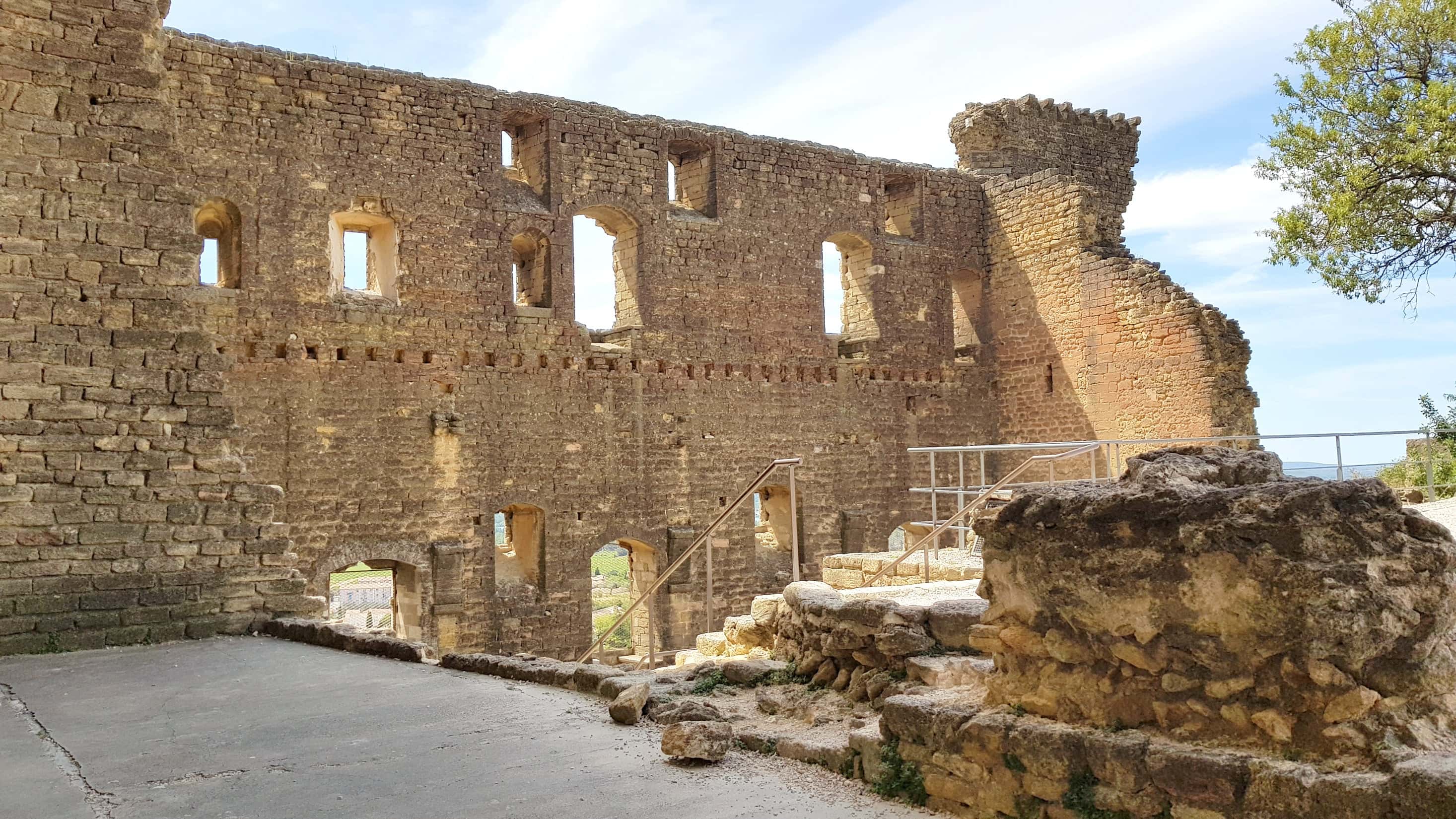 Ruins of an ancient stone castle with arched windows and partial walls, overlooking a scenic valley under a blue sky.