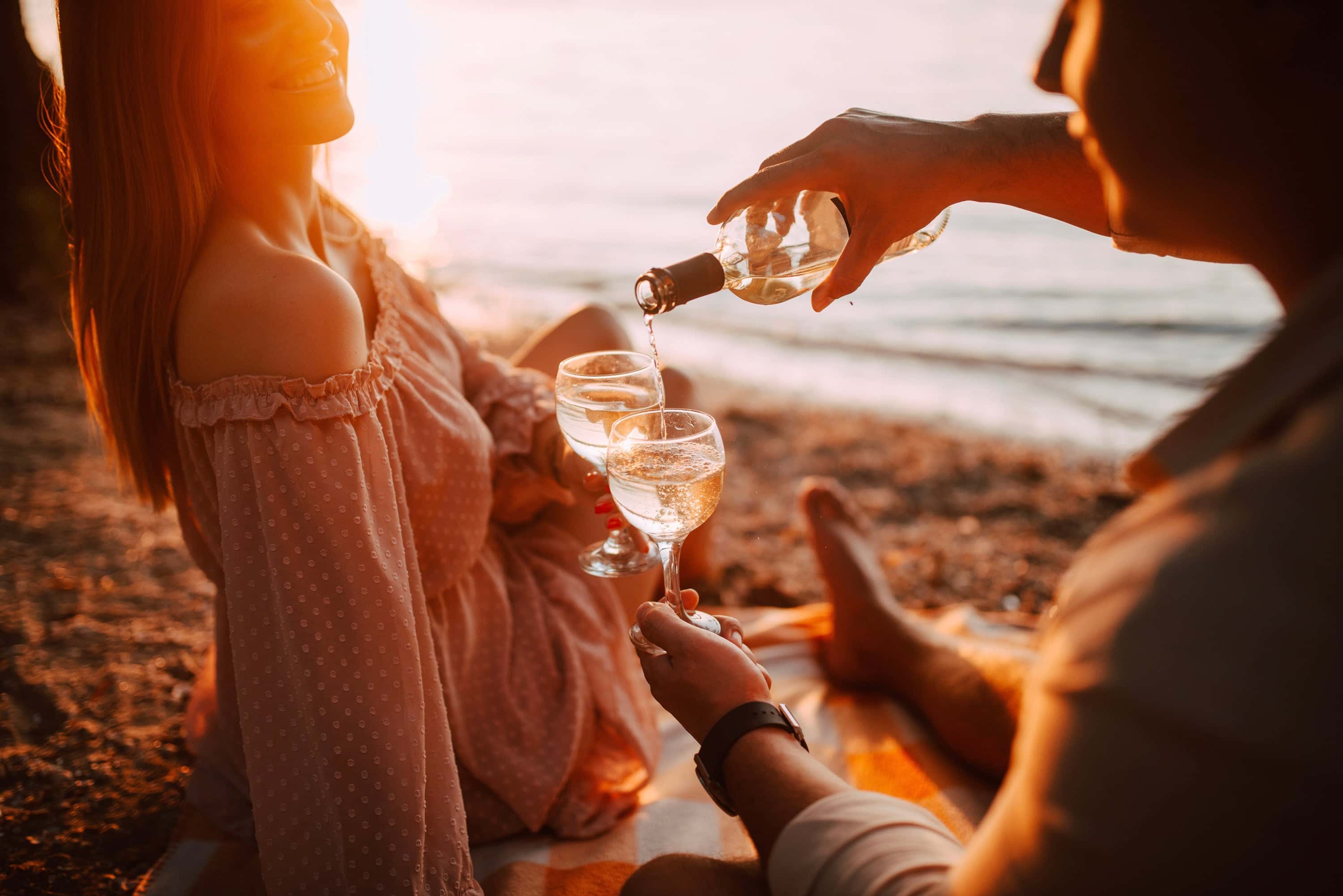 Romantic beach picnic at sunset with a couple pouring white wine into glasses while sitting on a blanket by the water.