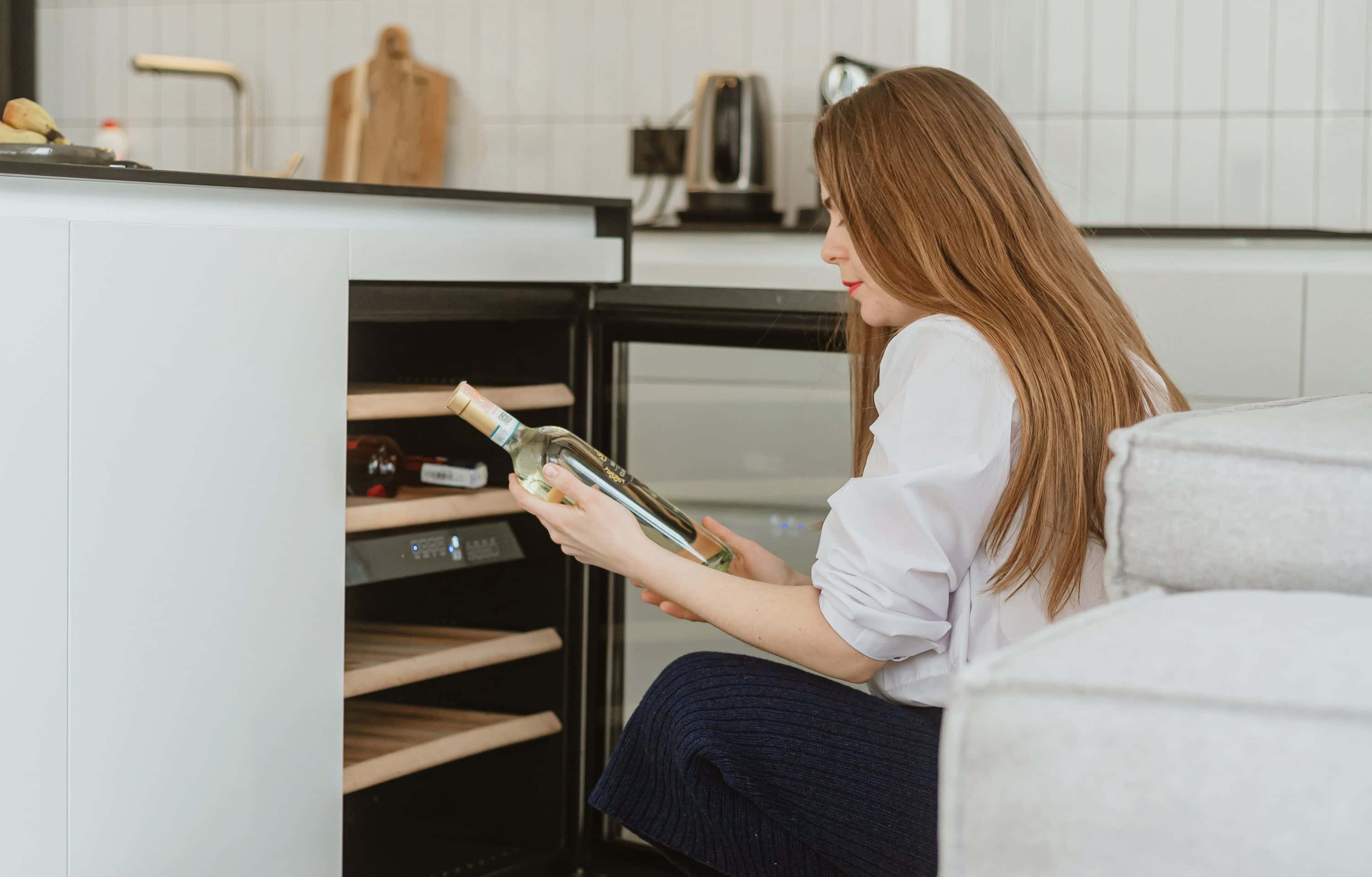 Woman selecting a bottle of white wine from a modern built-in wine fridge in a contemporary kitchen setting.