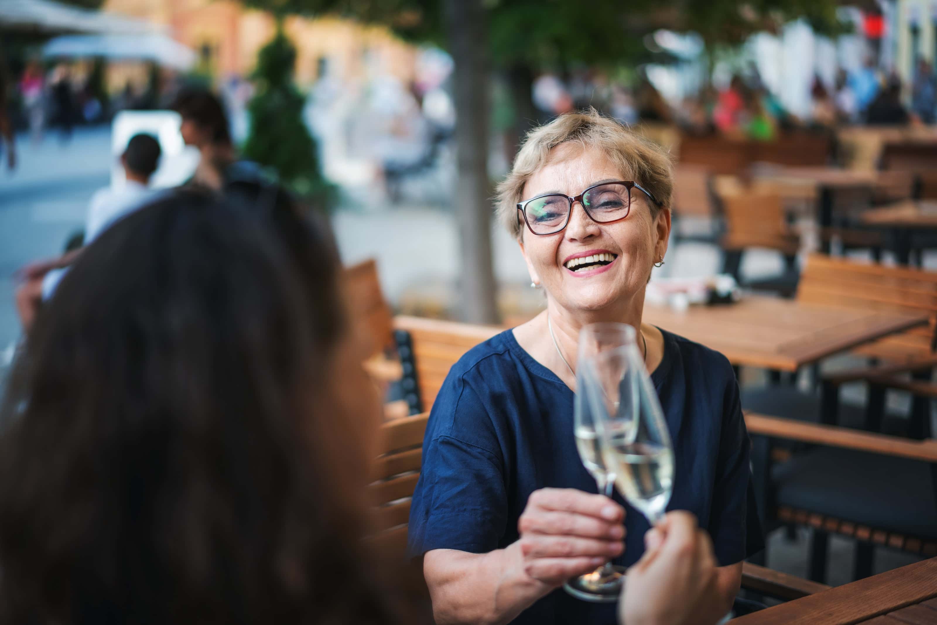 Smiling older woman clinking champagne glasses with a companion at an outdoor café, surrounded by wooden tables and a bustling city street.