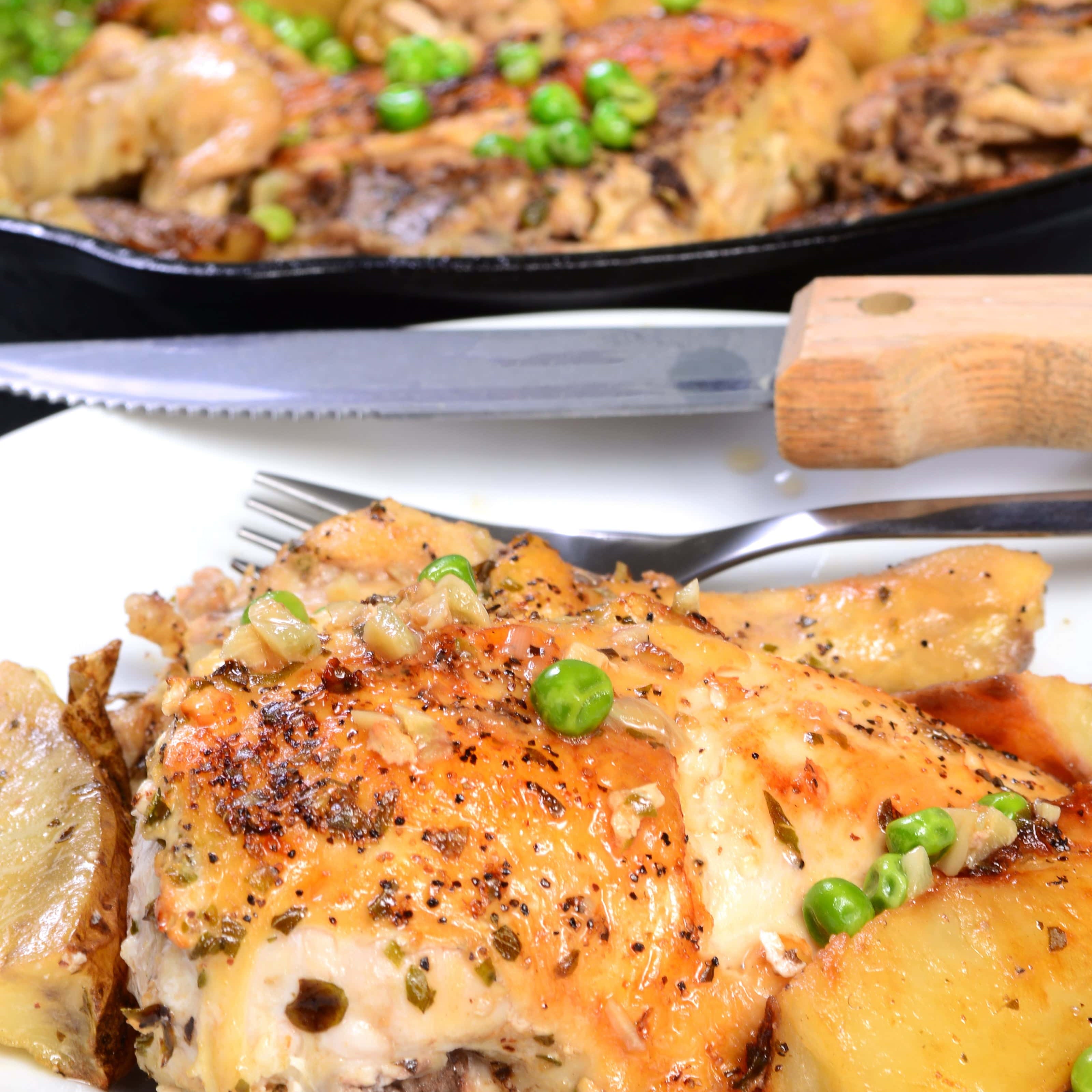 Close-up of a roasted chicken thigh served with golden-brown potatoes and green peas on a white plate, with a cast iron skillet filled with more of the dish in the background.