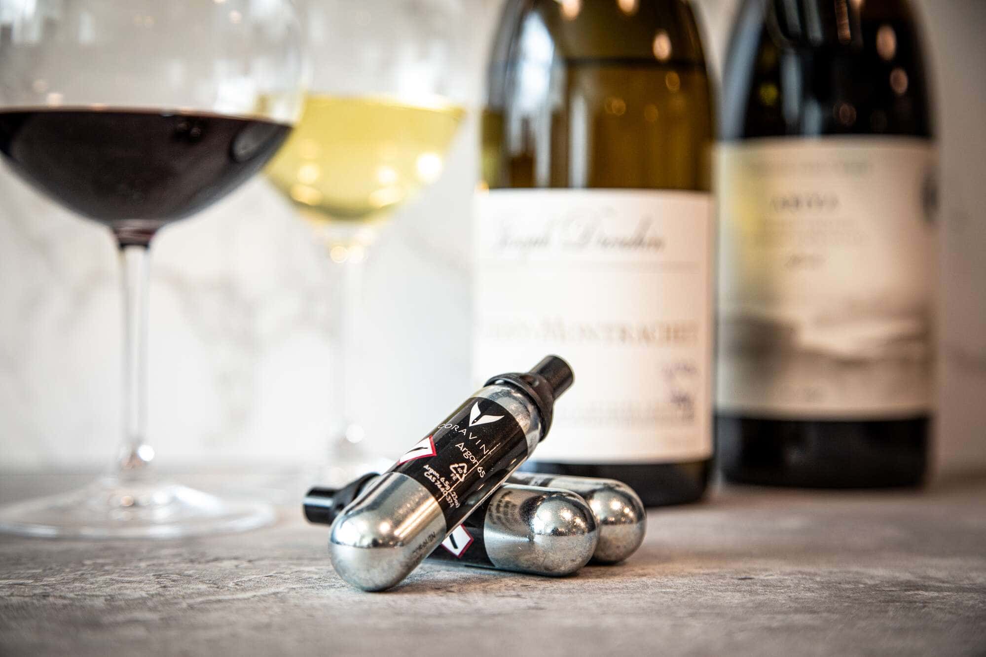 Close-up of Coravin argon gas capsules on a countertop with glasses of red and white wine and blurred wine bottles in the background.