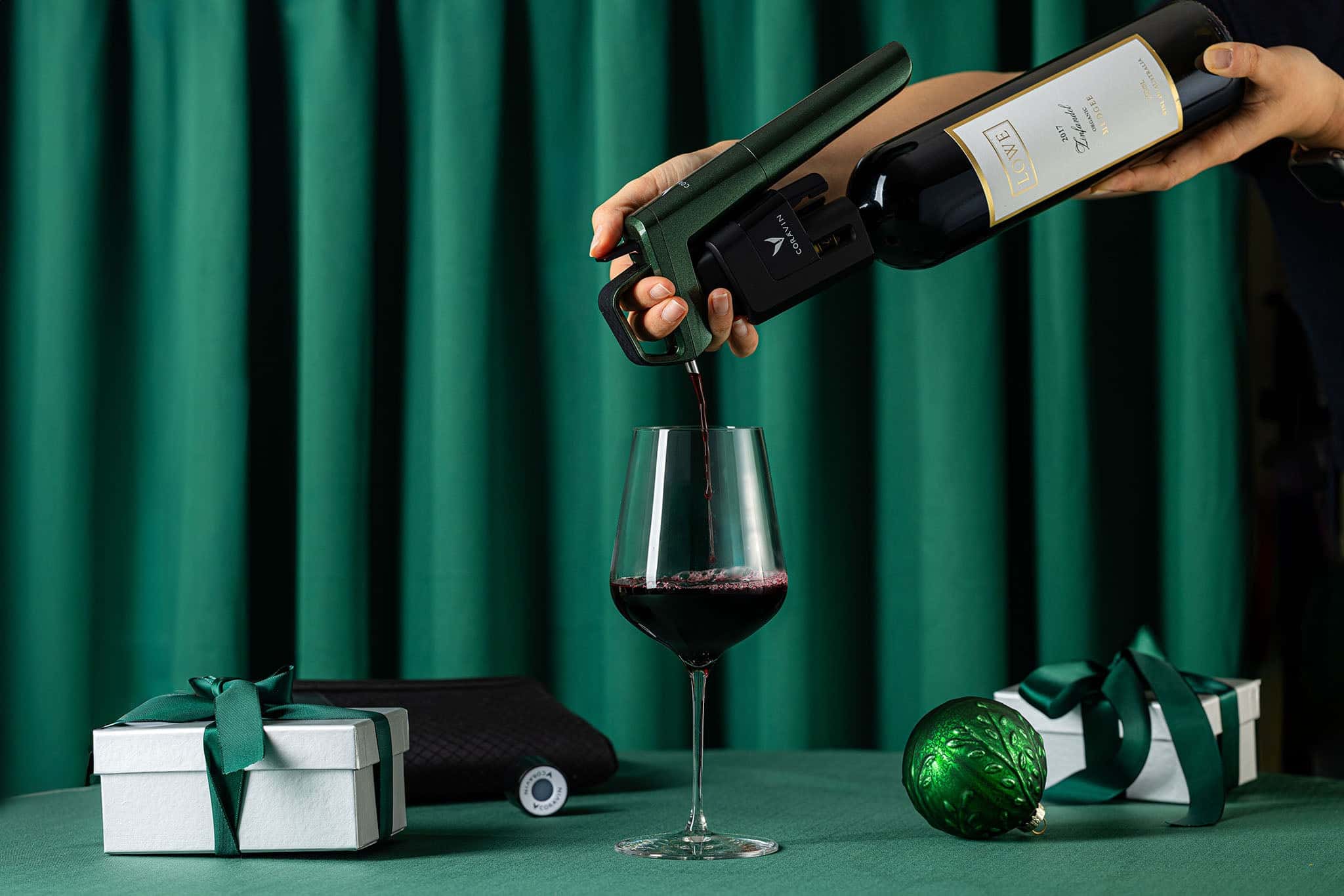 Hand using a Coravin wine preservation system to pour red wine into a glass, with green holiday gift boxes and an ornament on a green table and backdrop.