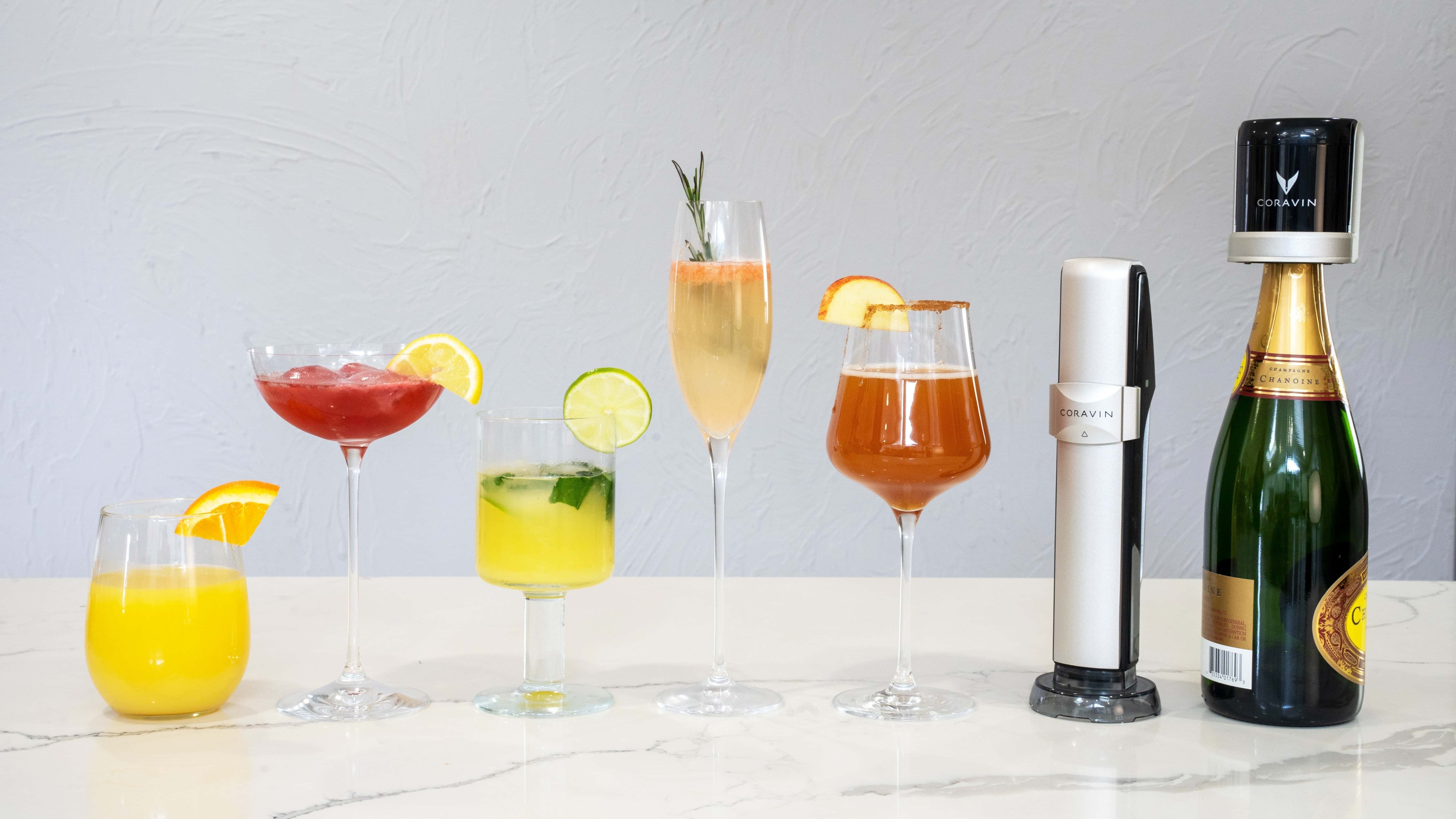 Assorted sparkling wine cocktails displayed in elegant glassware alongside a Coravin Sparkling system and champagne bottle on a marble countertop.