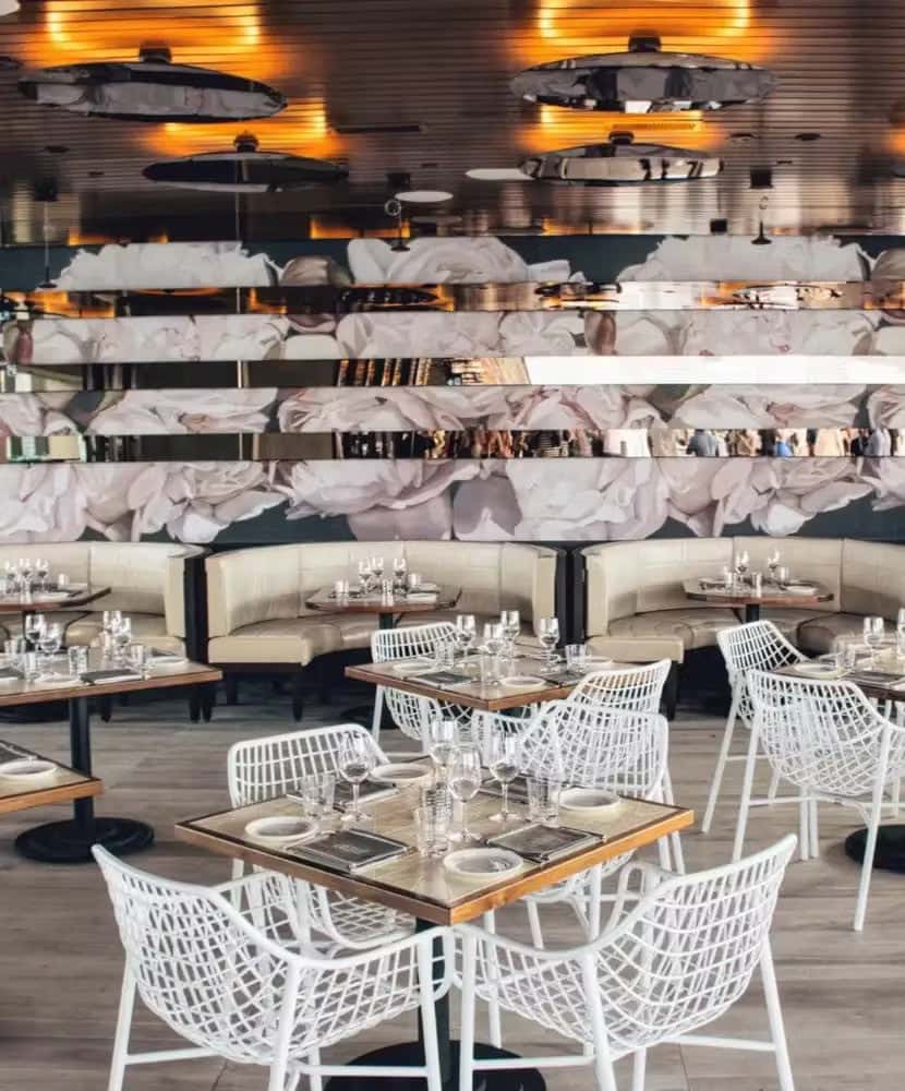 Chic indoor restaurant with modern white lattice chairs, wood-trimmed tables set for dining, curved beige booths, and a large floral mural accented by mirrored horizontal panels on the back wall.
