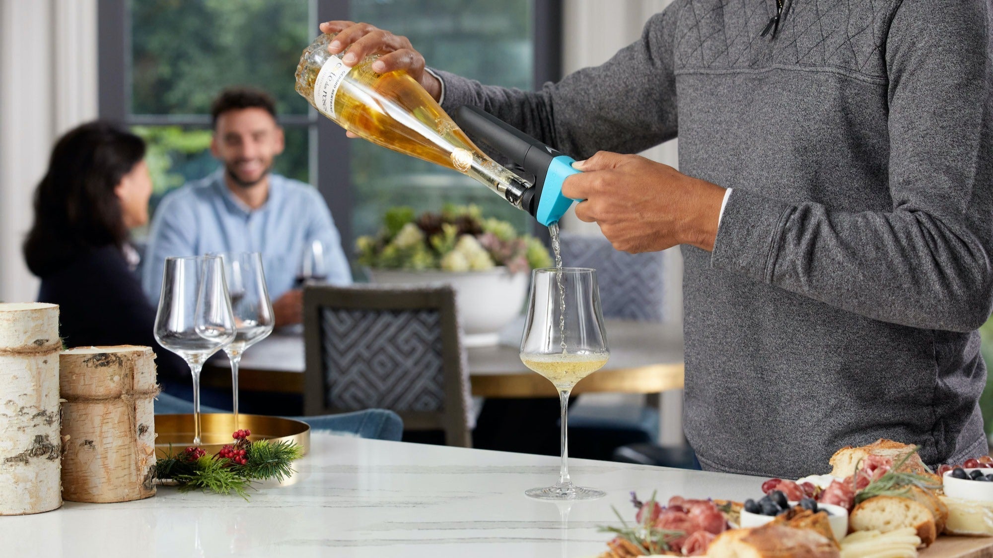 Person using a Coravin Pivot device to pour white wine into a glass, with a charcuterie board in the foreground and two people conversing in the background at a modern, well-lit dining space.