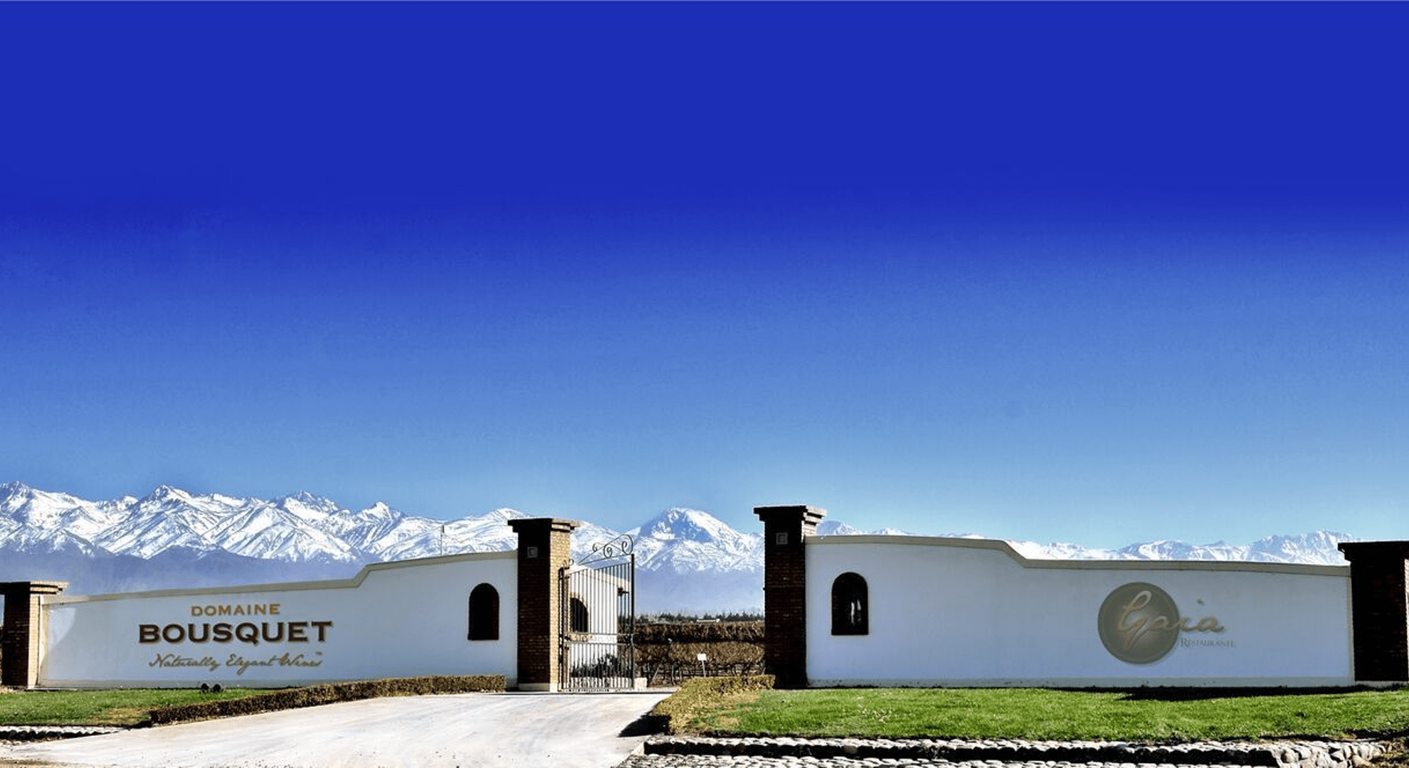 Entrance gate to Domaine Bousquet winery with snow-capped Andes mountains in the background under a vivid blue sky.