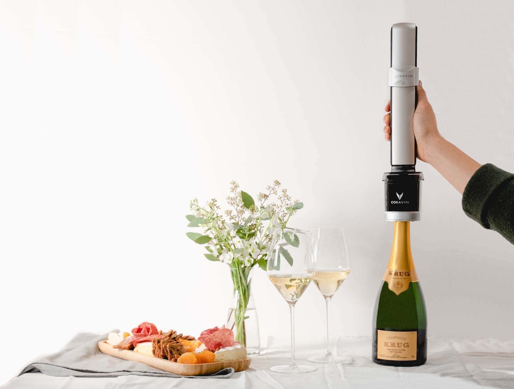 A hand using a Coravin Sparkling wine preservation system on a bottle of Krug Champagne, placed beside two filled wine glasses, a charcuterie board with meats and cheeses, and a small vase of white flowers on a white tablecloth.