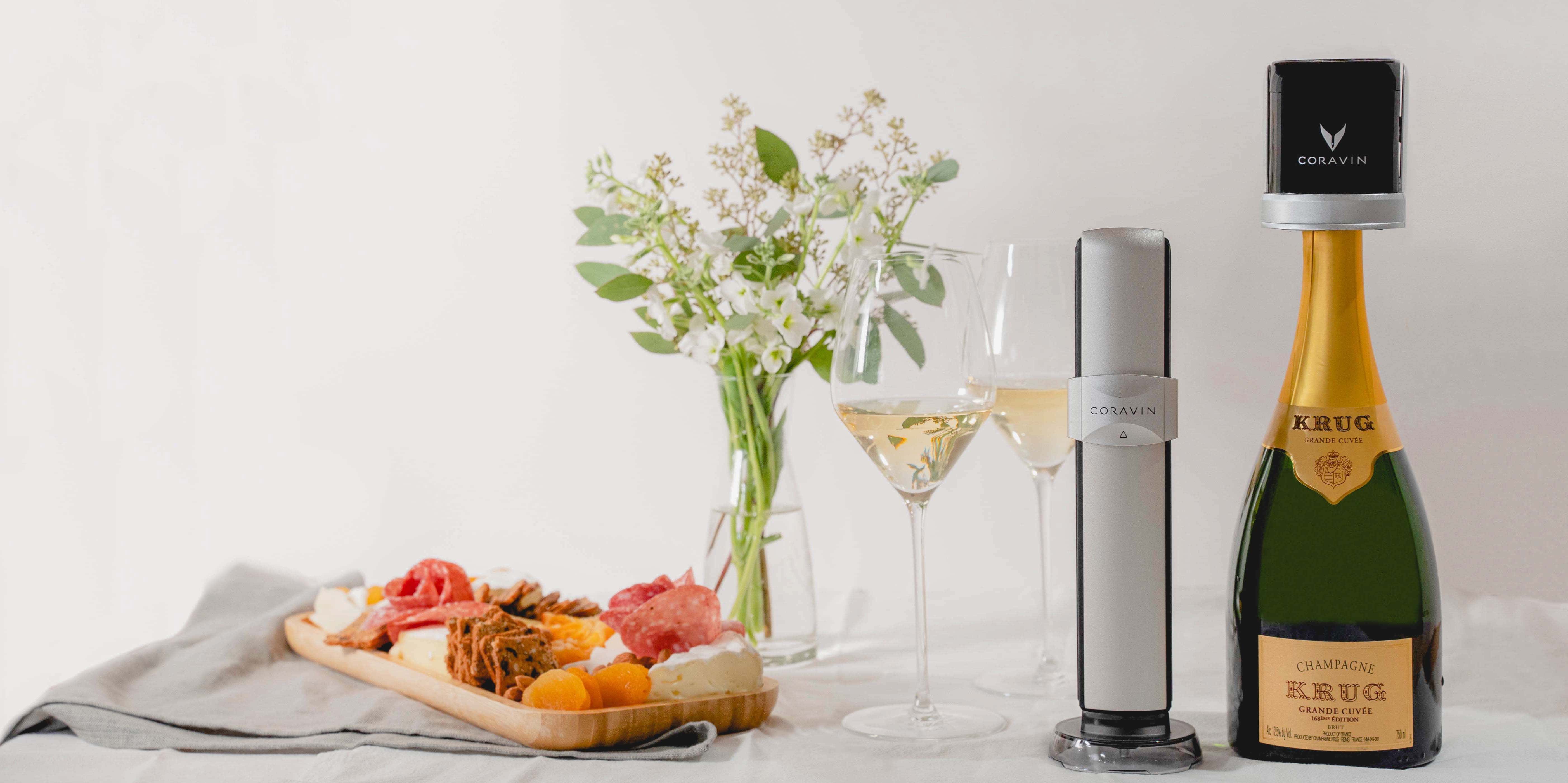 Coravin Sparkling system attached to a bottle of Krug Champagne, with two filled glasses, a charcuterie board, and a vase of flowers on a white tablecloth.