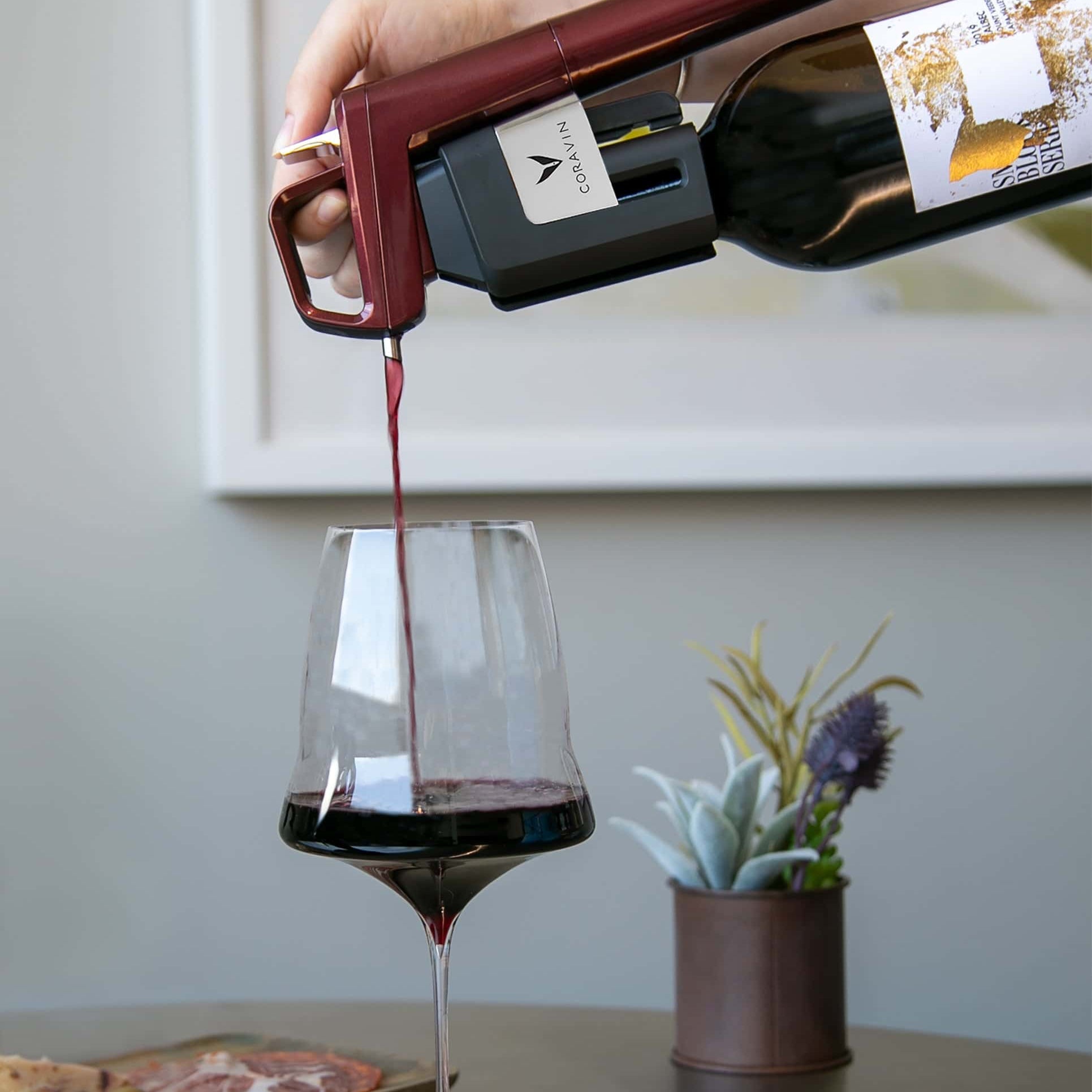 Red wine being poured into a glass using a Coravin wine preservation system, with a charcuterie board and small potted plant in the background.