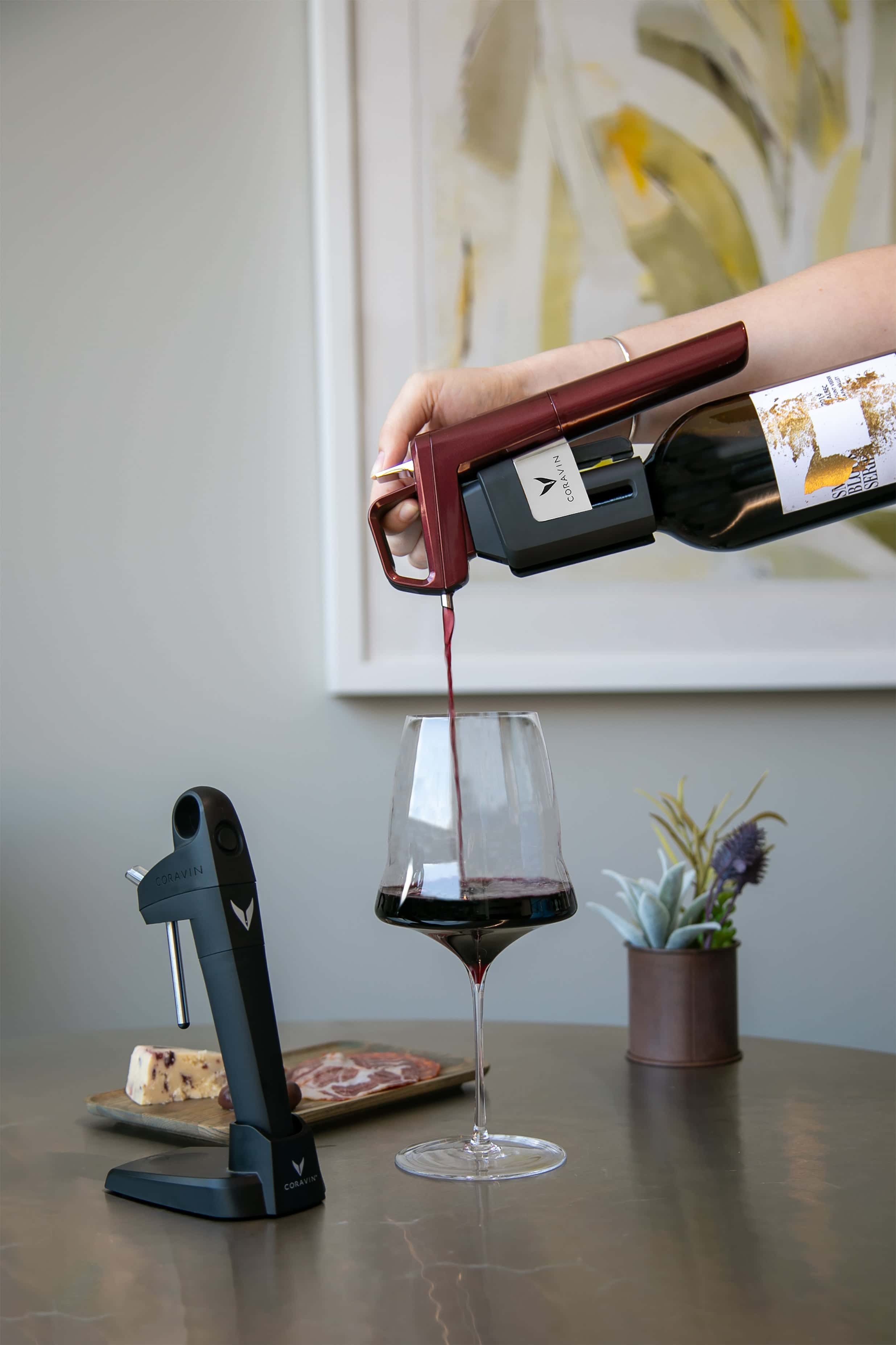 Hand pouring red wine from a bottle using a Coravin Timeless wine preservation system into a glass, with a Coravin Pivot device, charcuterie, and cheese on the table in the background.