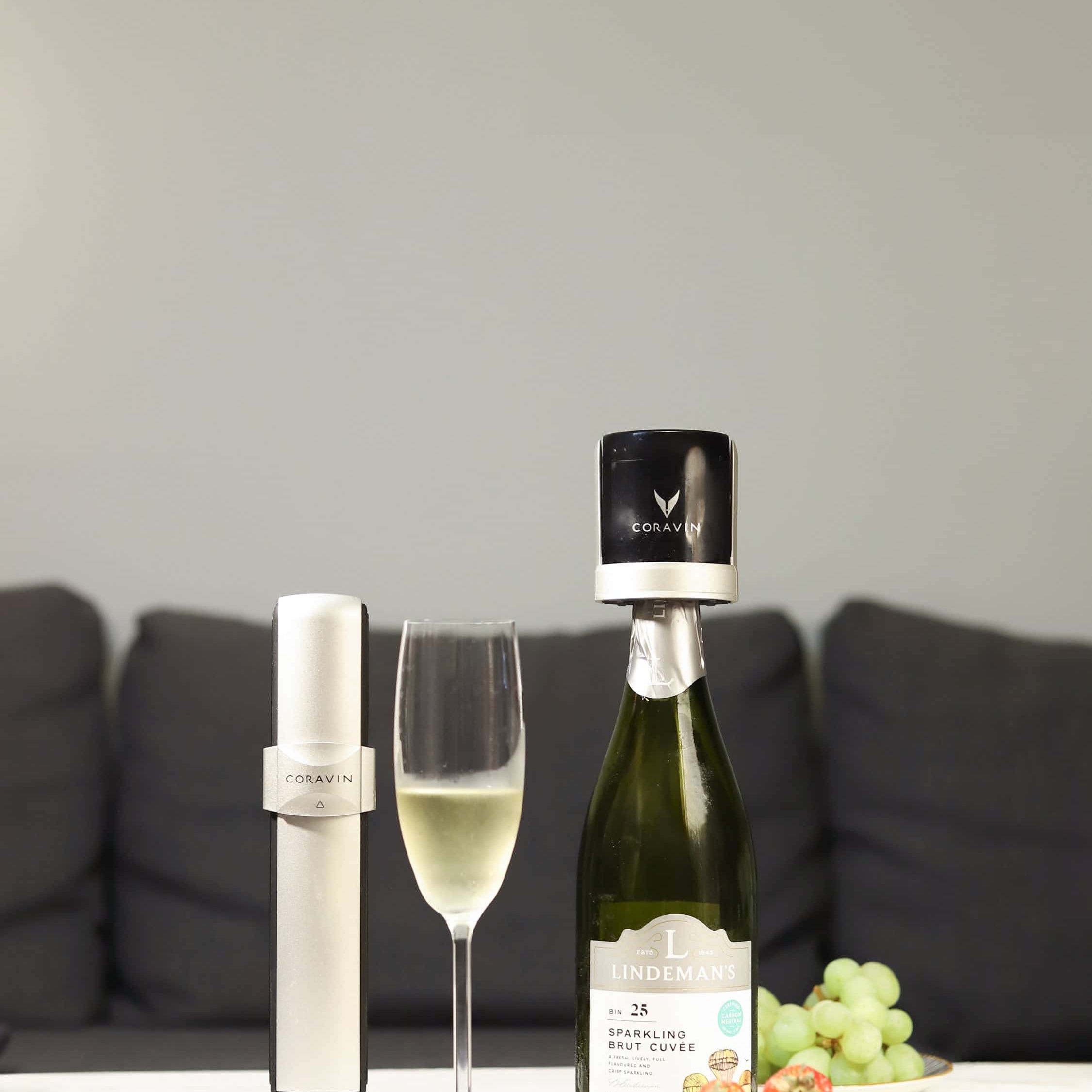 Coravin Sparkling wine preservation system displayed beside a bottle of Lindeman’s sparkling brut cuvée with a Coravin stopper, a filled champagne flute, and plates of fresh strawberries and green grapes on a white table in front of a dark sofa.