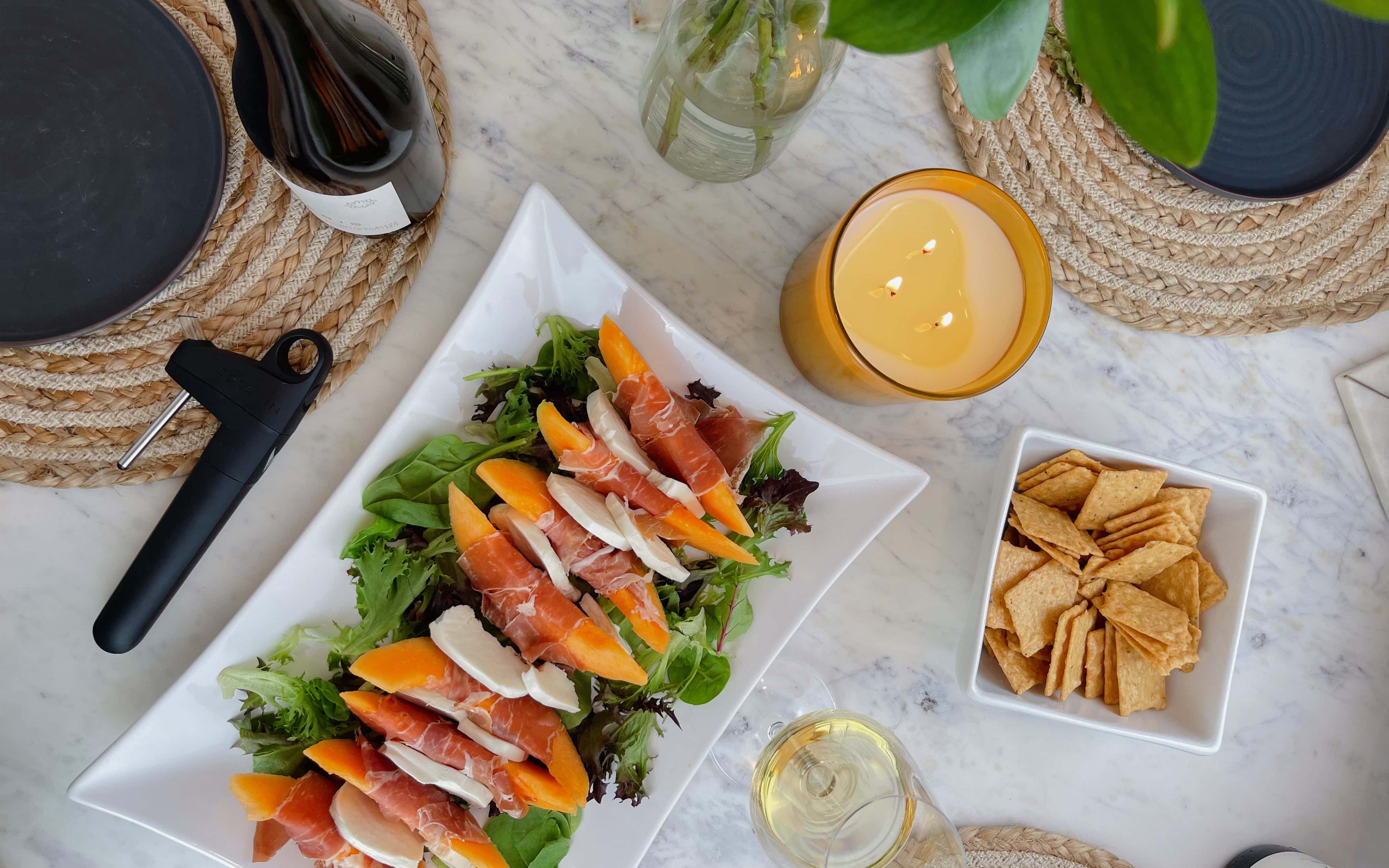 Overhead view of a summer table setting with a plate of prosciutto-wrapped melon and mozzarella on greens, crackers, a glass of white wine, a lit candle, and a Coravin wine preservation system.
