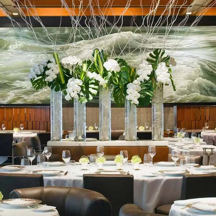 Elegant fine dining restaurant interior with round tables set with white tablecloths, glassware, and floral centerpieces, featuring tall glass vases with white orchids and large green leaves in the center of the room.